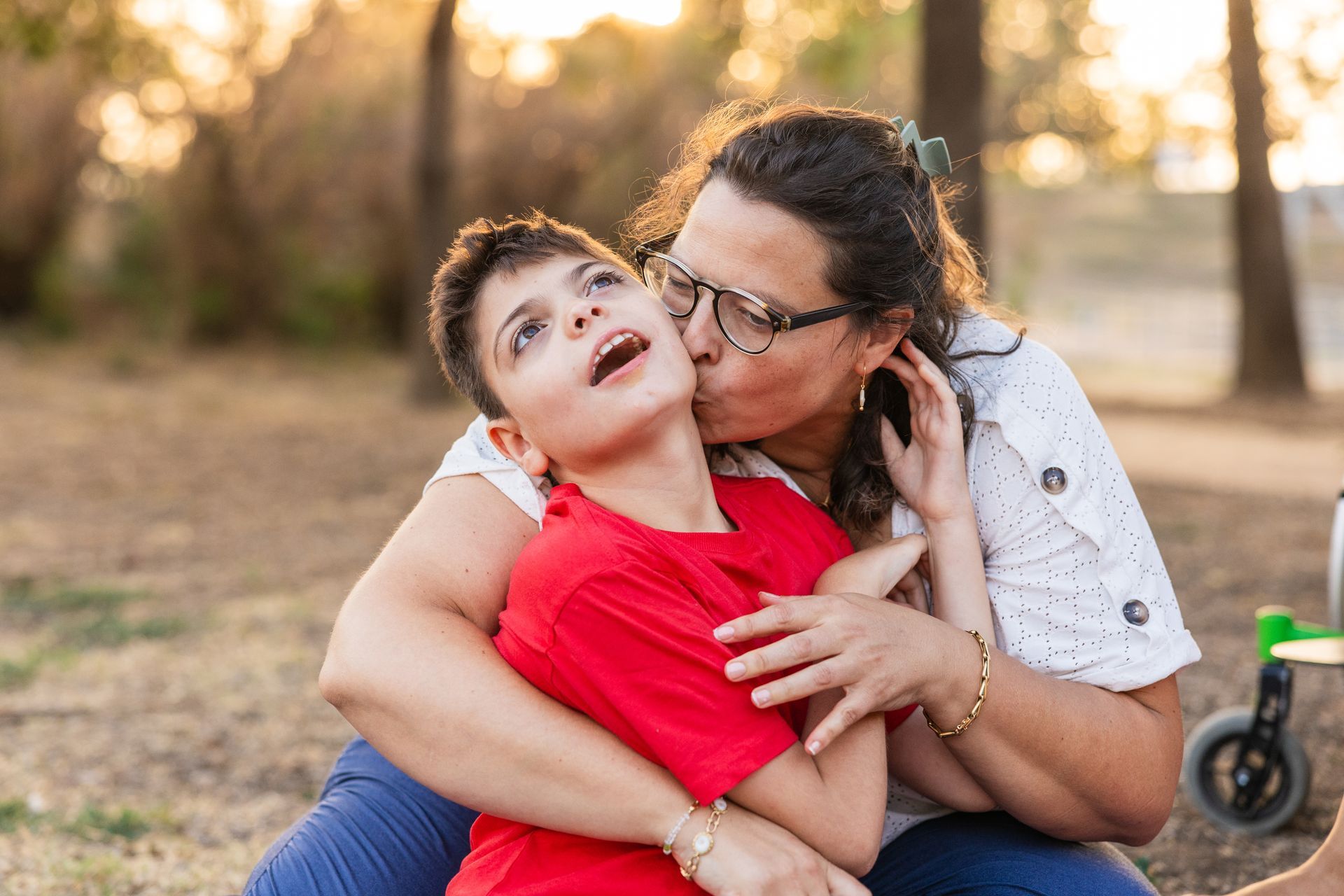 Adult kissing child on the cheek outdoors, both seated on grass in warm sunlight
