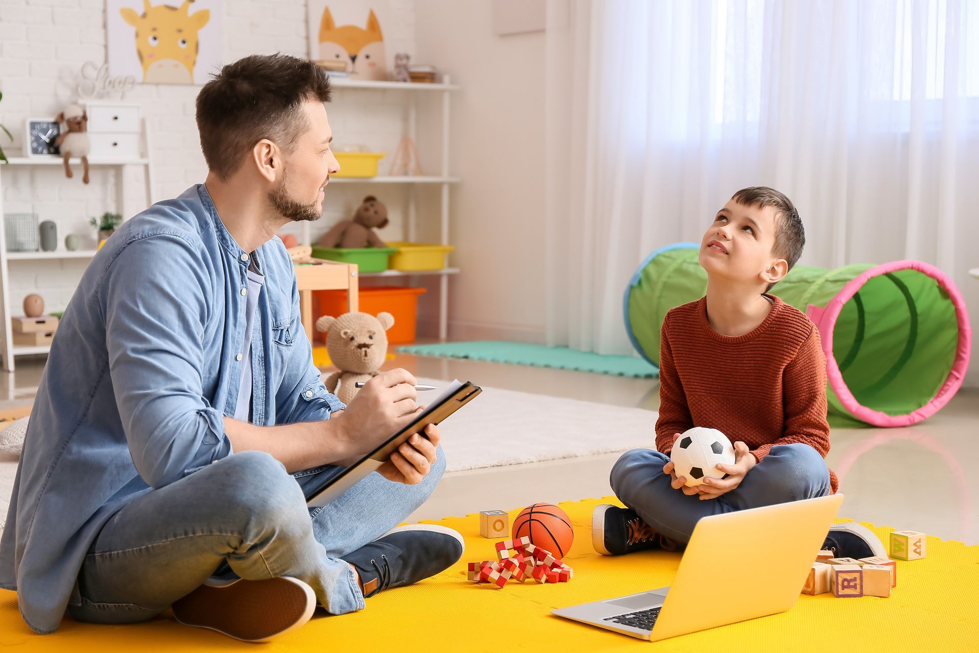 Adult and child sitting on the floor in a bright playroom, talking and playing with toys and a laptop.
