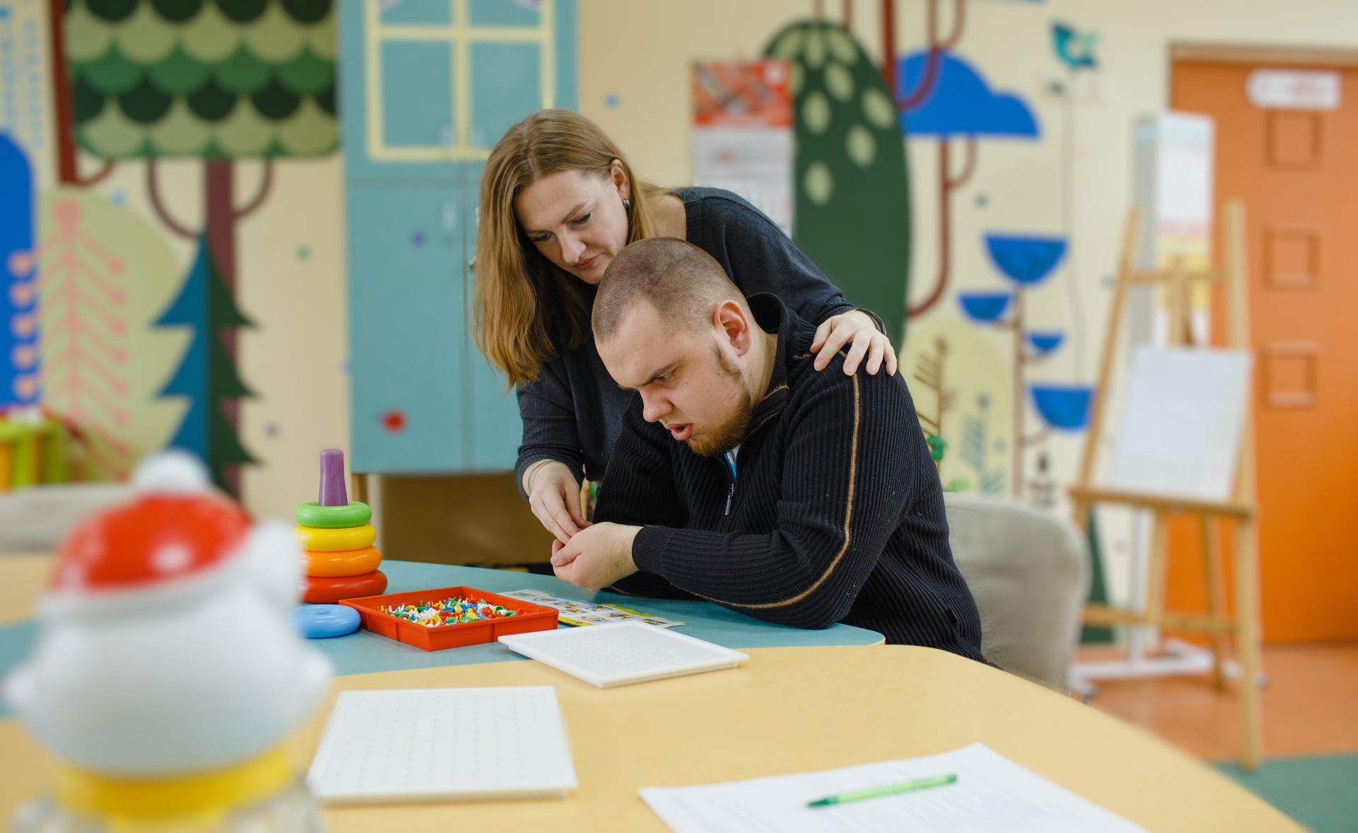 Two people leaning over a table in a colorful classroom, focused on a project.