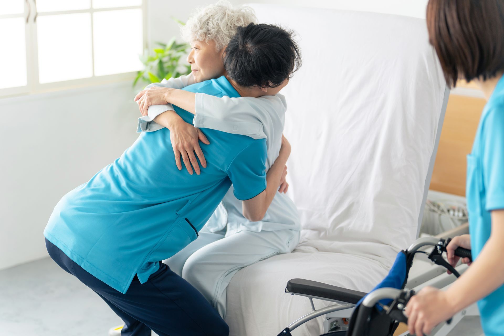 Caregiver in blue shirt hugs seated patient beside hospital bed, with wheelchair nearby