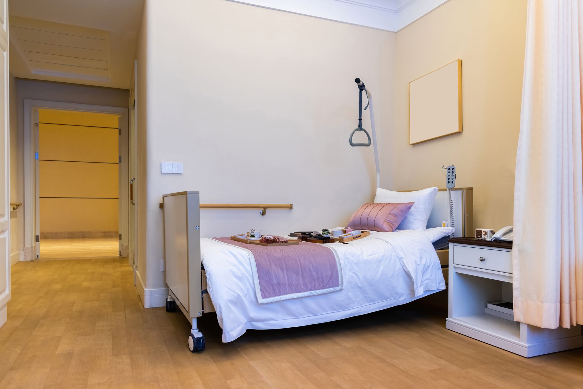 Small hospital room with a single bed, bedside table, wooden floor, and beige walls
