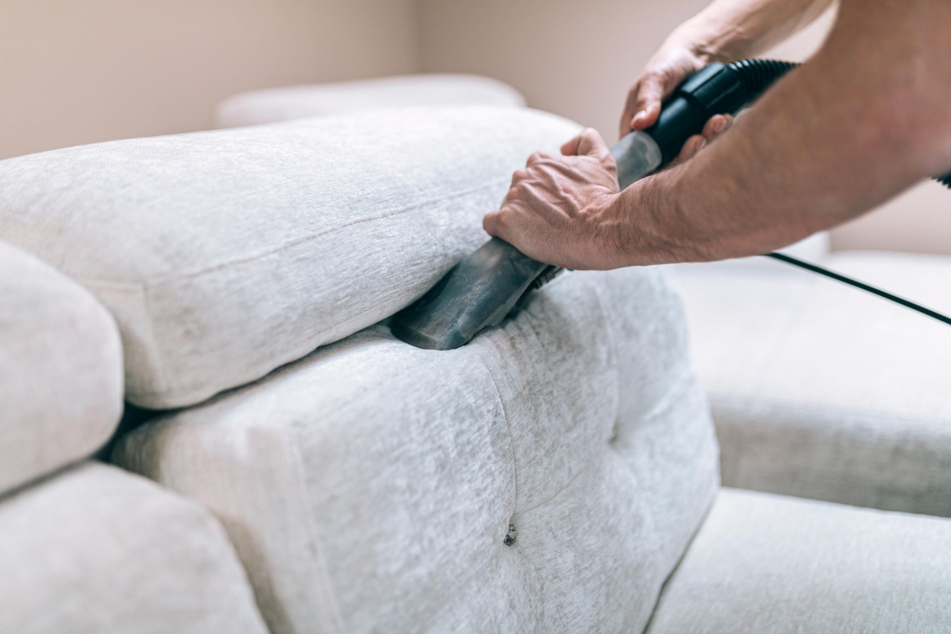Person vacuuming a white couch with a handheld vacuum cleaner.