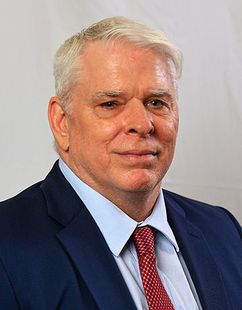 A man in a blue suit, light blue dress shirt, and red patterned tie against a plain white background.