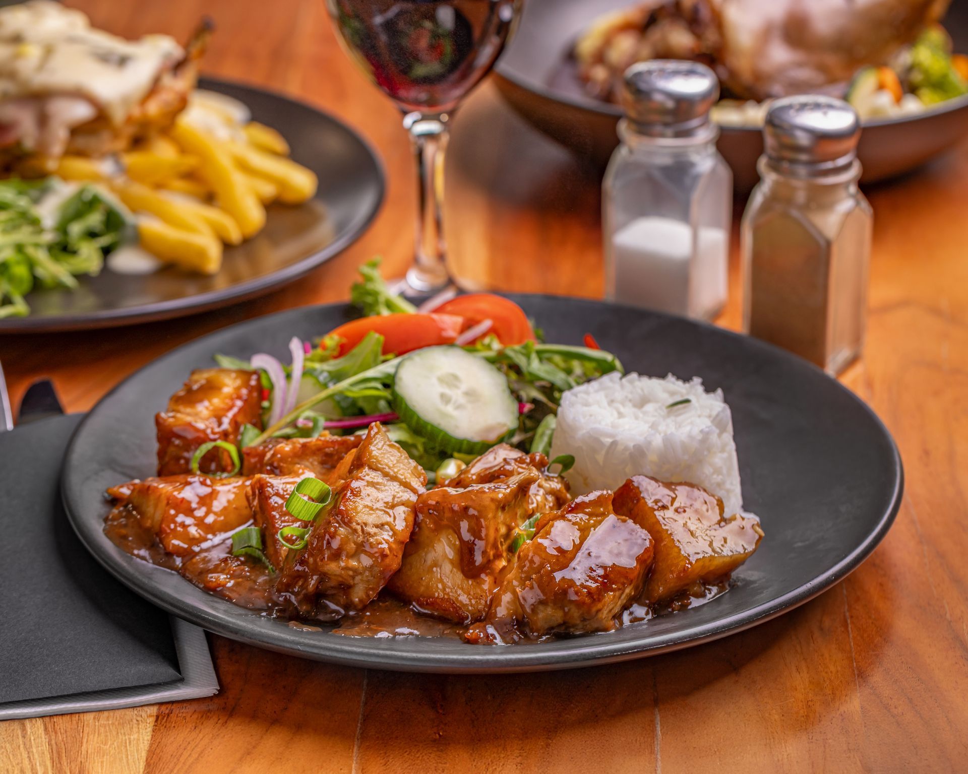 Pork dish with rice, salad, and sauce on a dark plate. Red wine and other dishes in background — Inverell Sporties in Inverell, NSW
