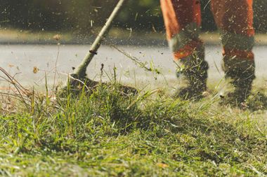 Person using a string trimmer sprays wet grass near a road