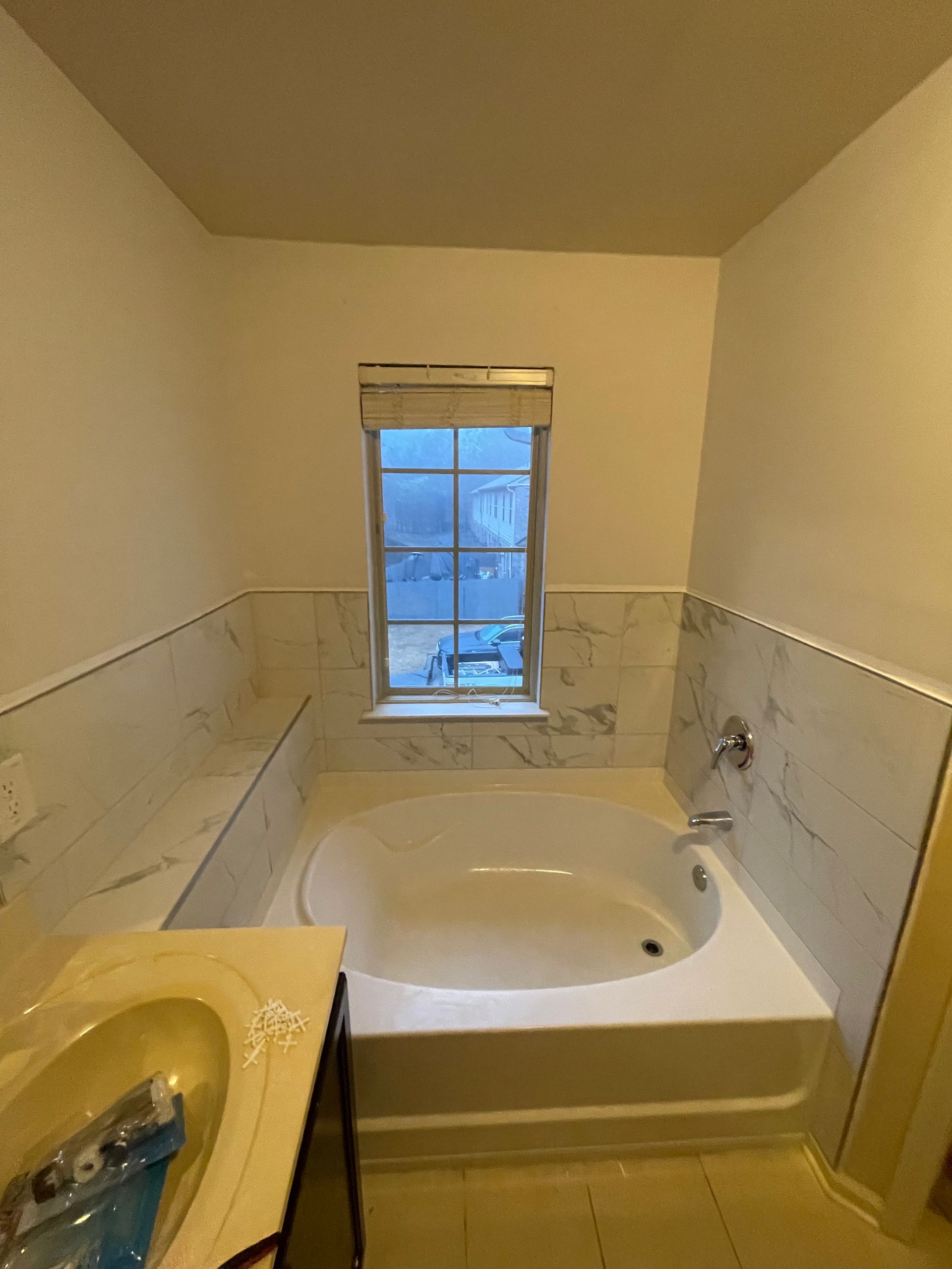 Bathroom with white tub, marble tile, and a window with blinds.