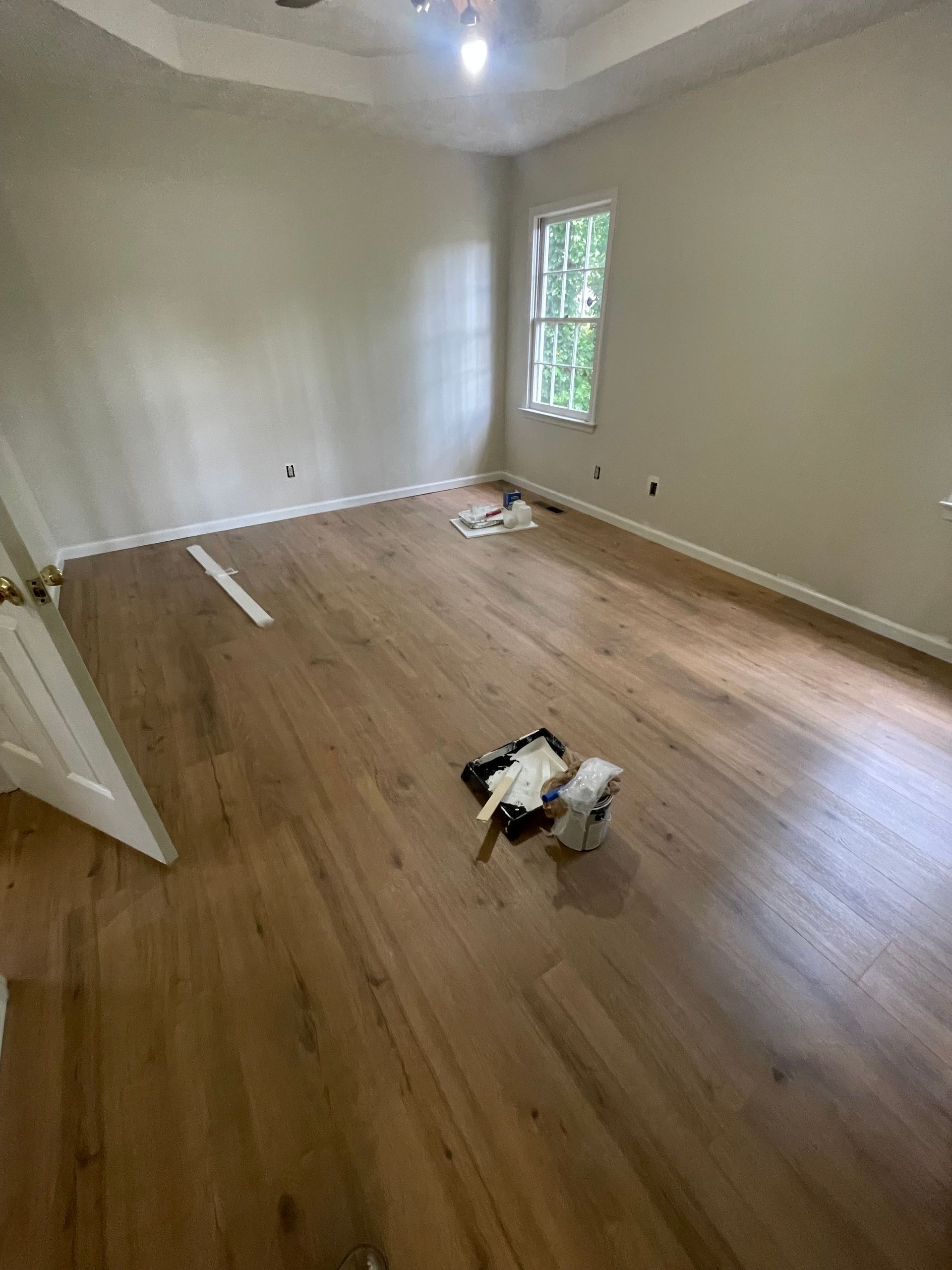 Empty room with hardwood floors, beige walls, and a small window. Paint supplies are on the floor.