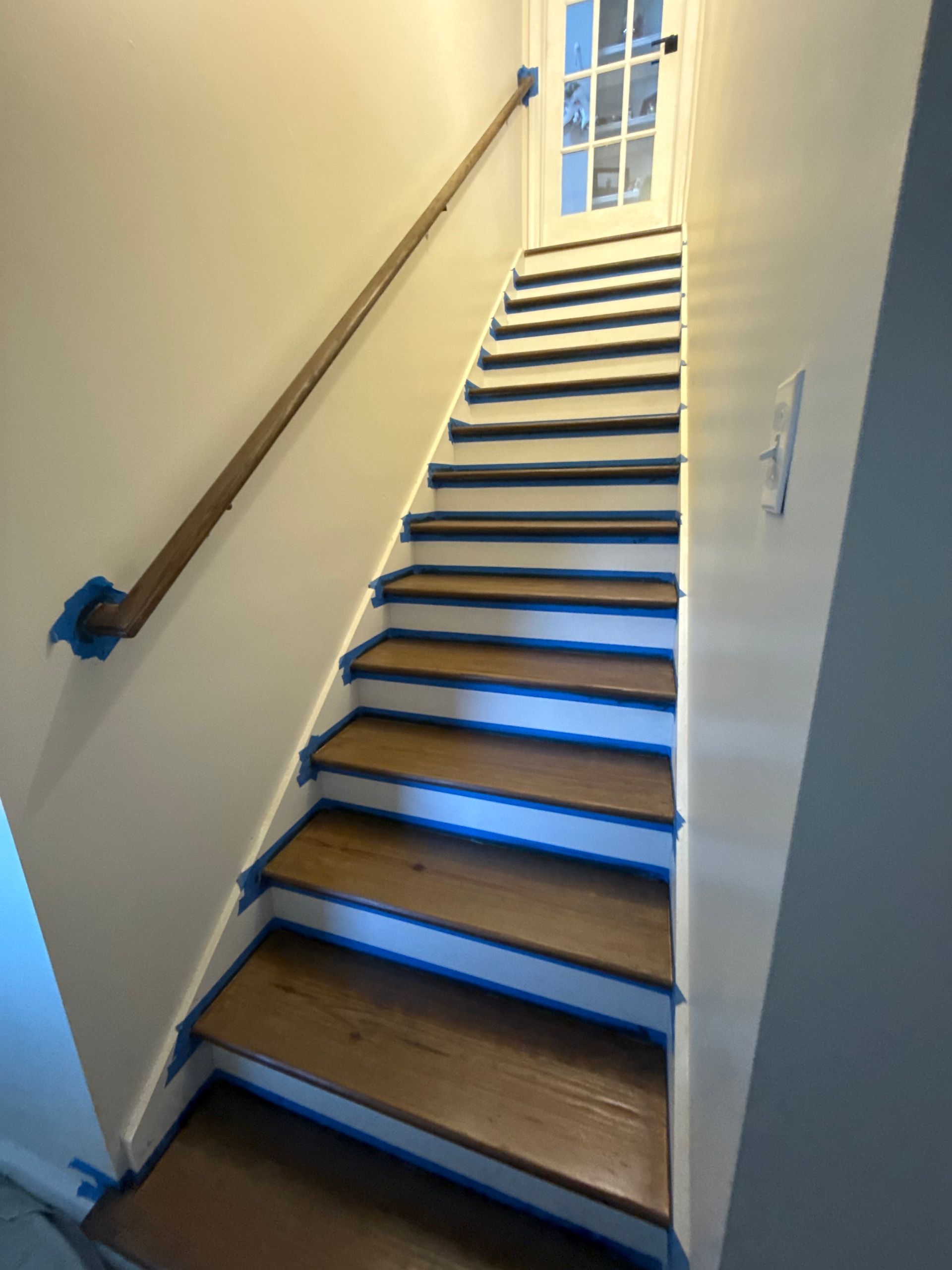 Staircase with wooden steps and handrail, white walls, window at top, blue painter's tape along edges.