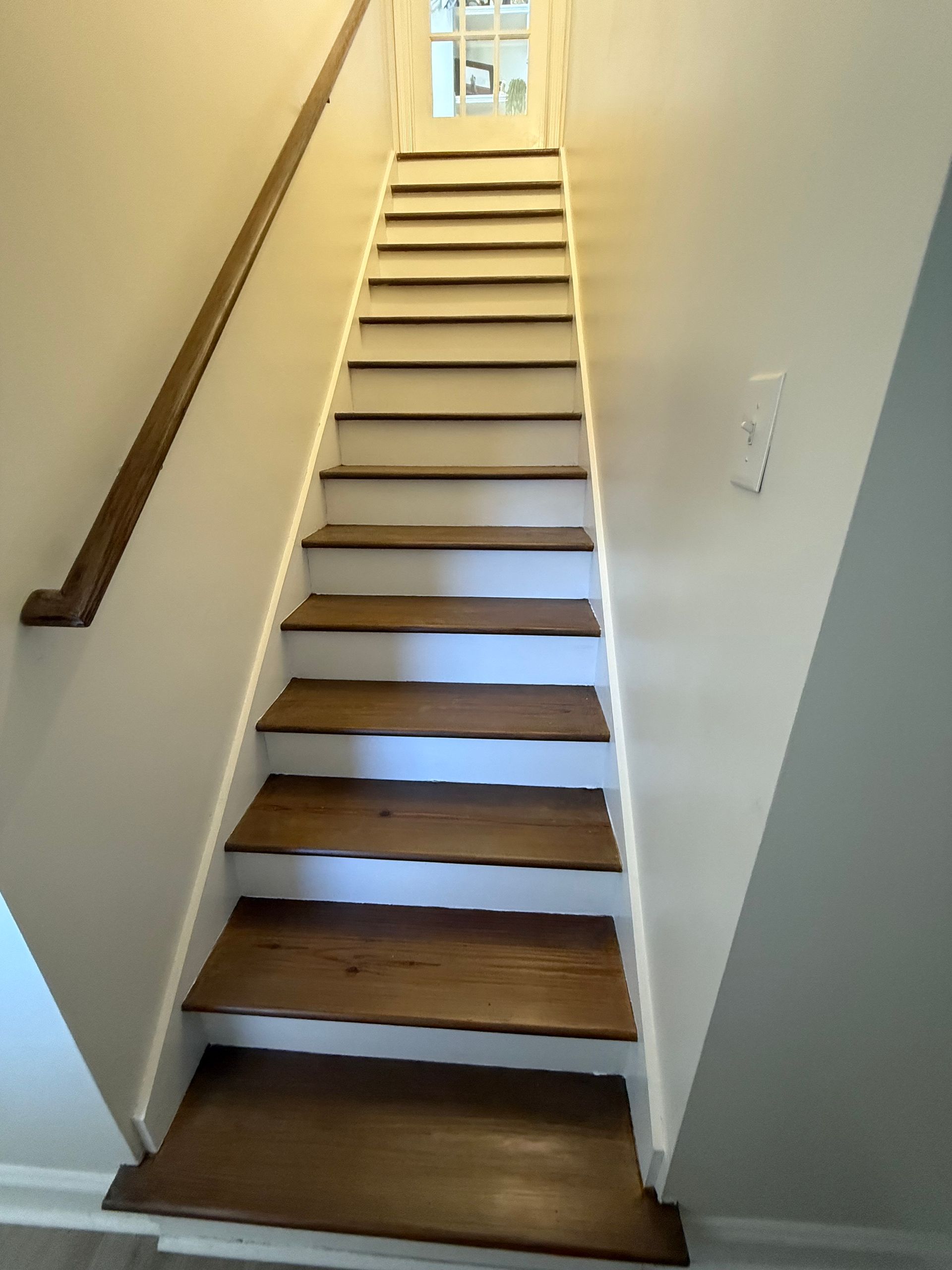 Wooden staircase with brown treads and white risers, leading upwards with a wooden handrail on the left.