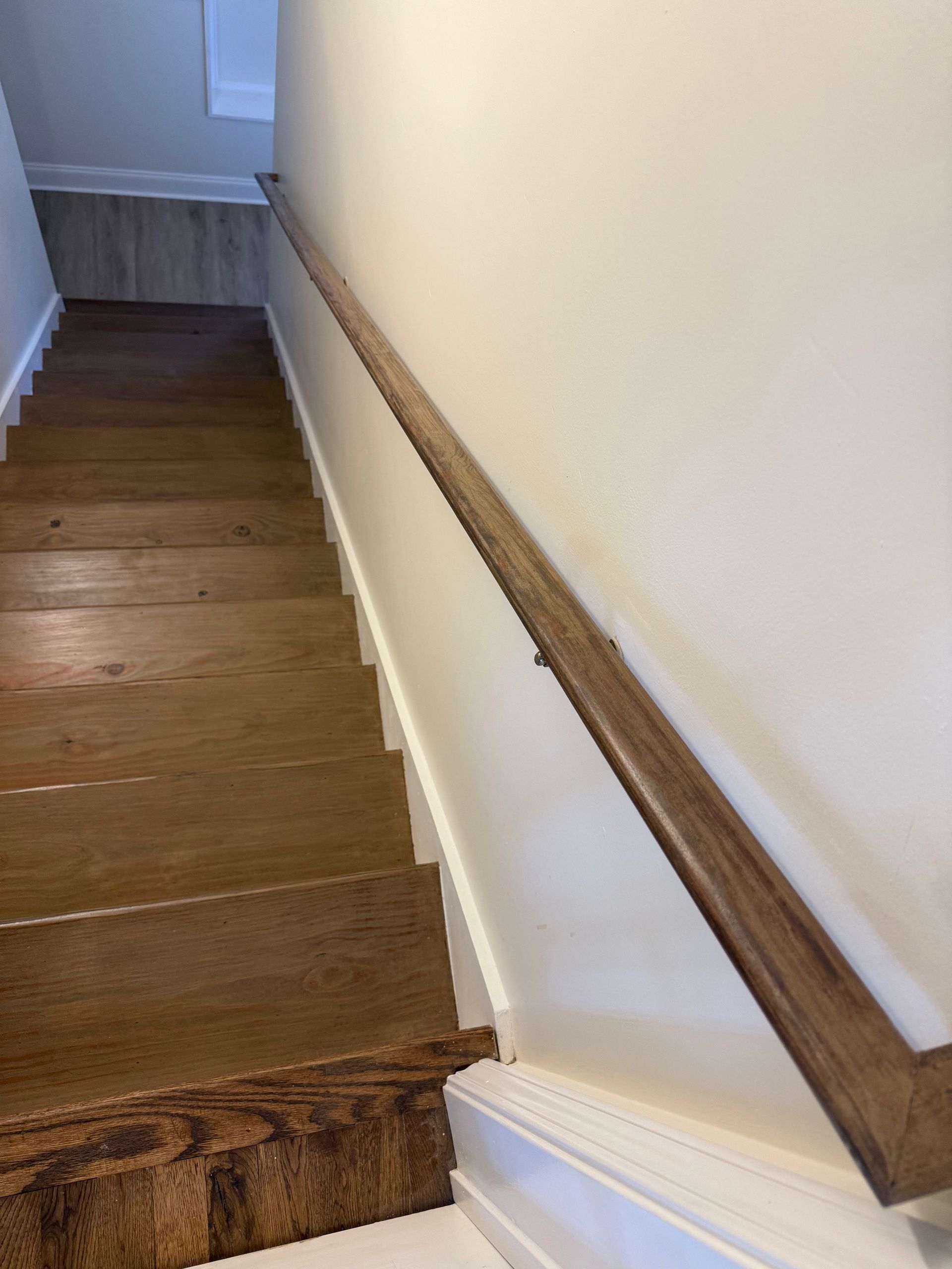 Wooden staircase with a dark wood handrail along a white wall.