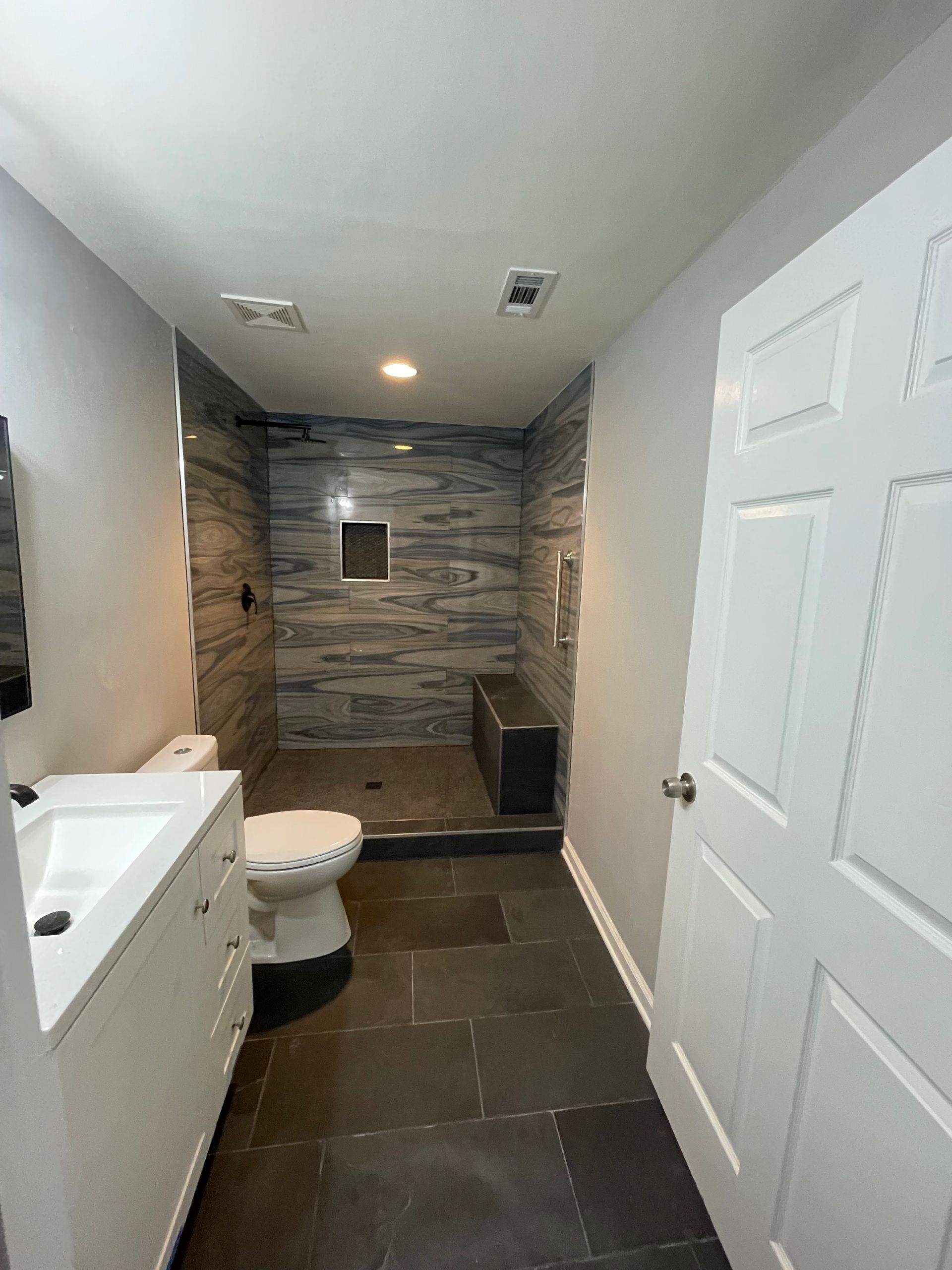 Bathroom with a gray and blue tiled shower, a white vanity and toilet, and a dark tile floor.