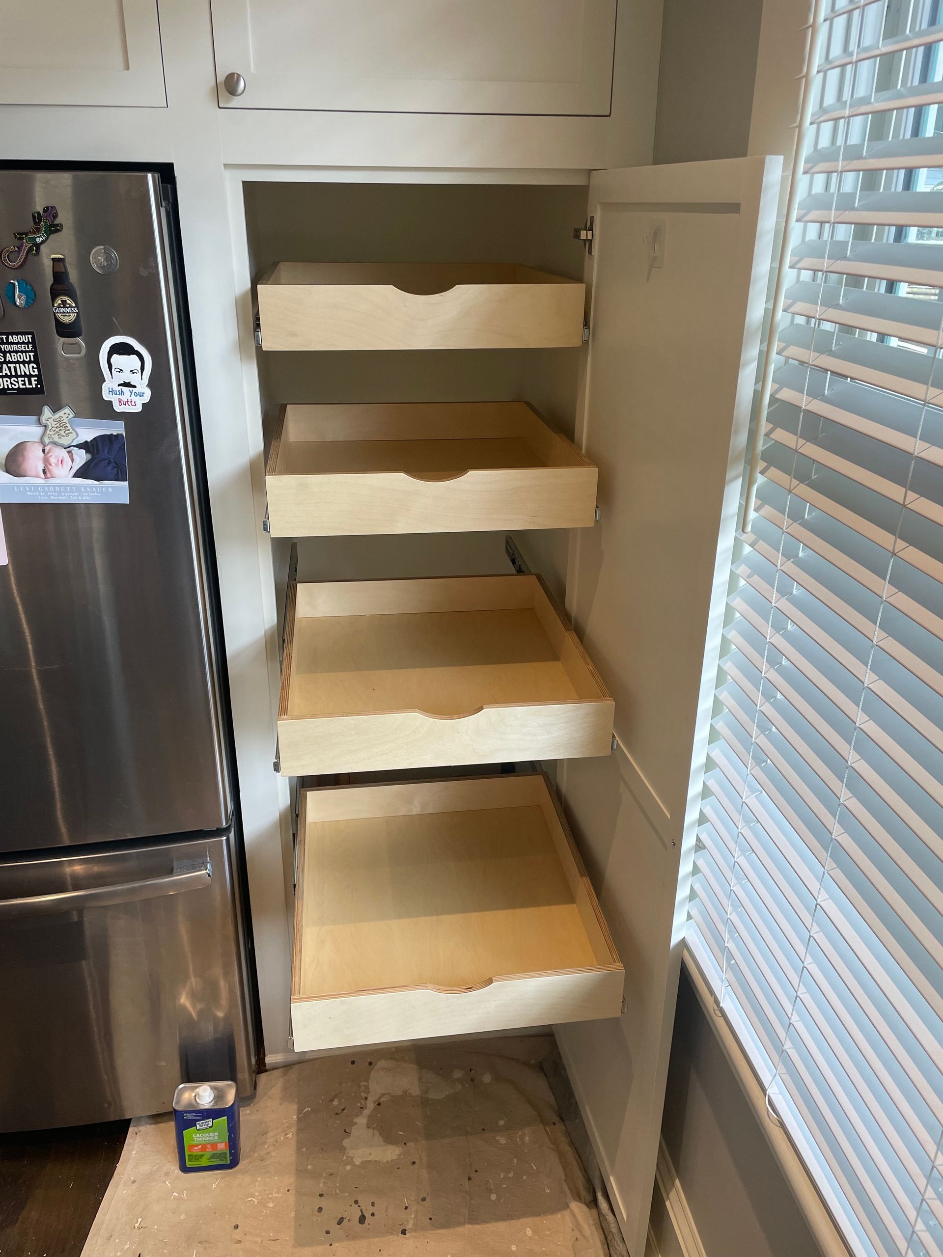 Wooden pull-out shelves inside a kitchen cabinet, next to a stainless steel refrigerator and window with blinds.