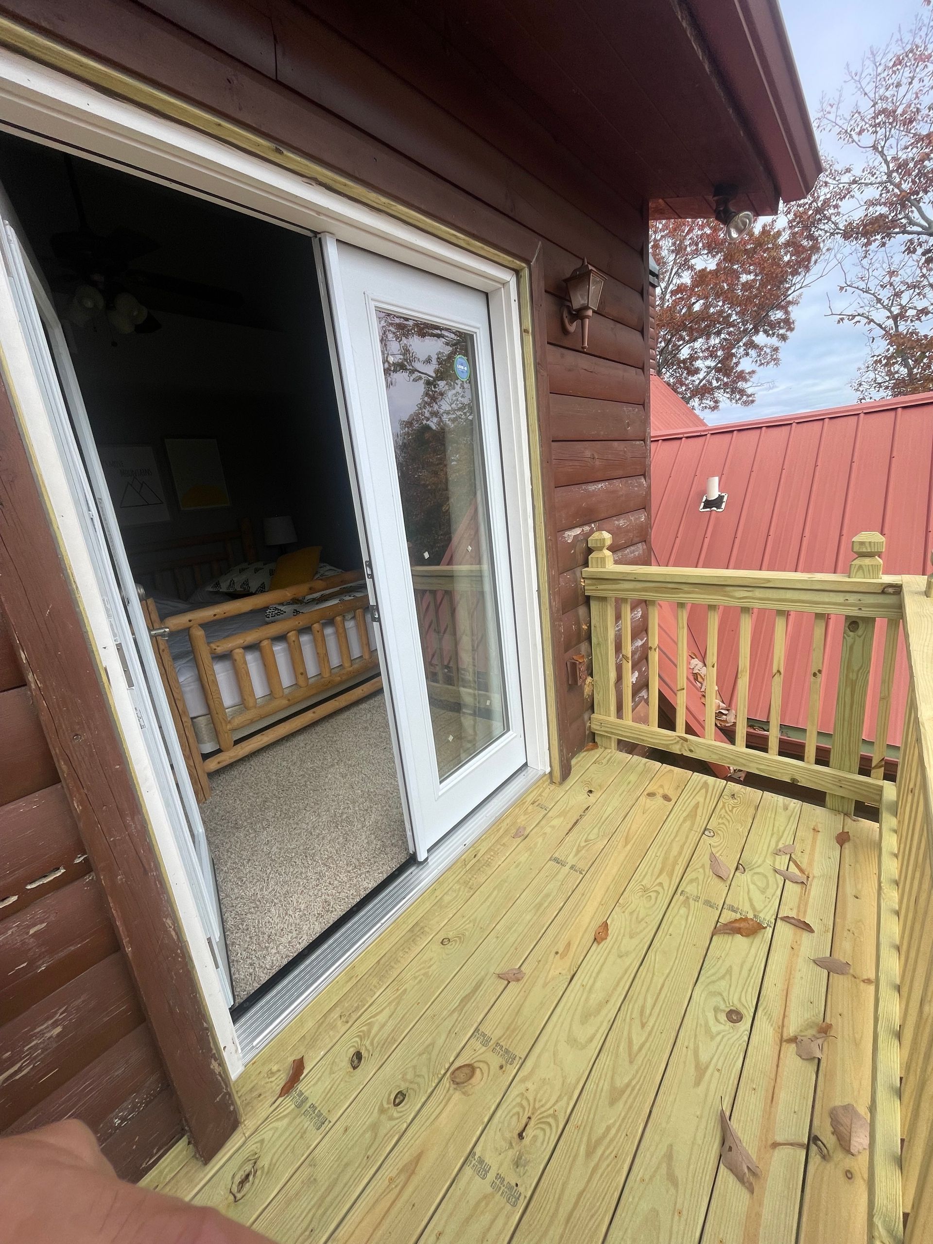 Deck with railing, open sliding door to bedroom.  Wooden deck, log siding, red metal roof visible.