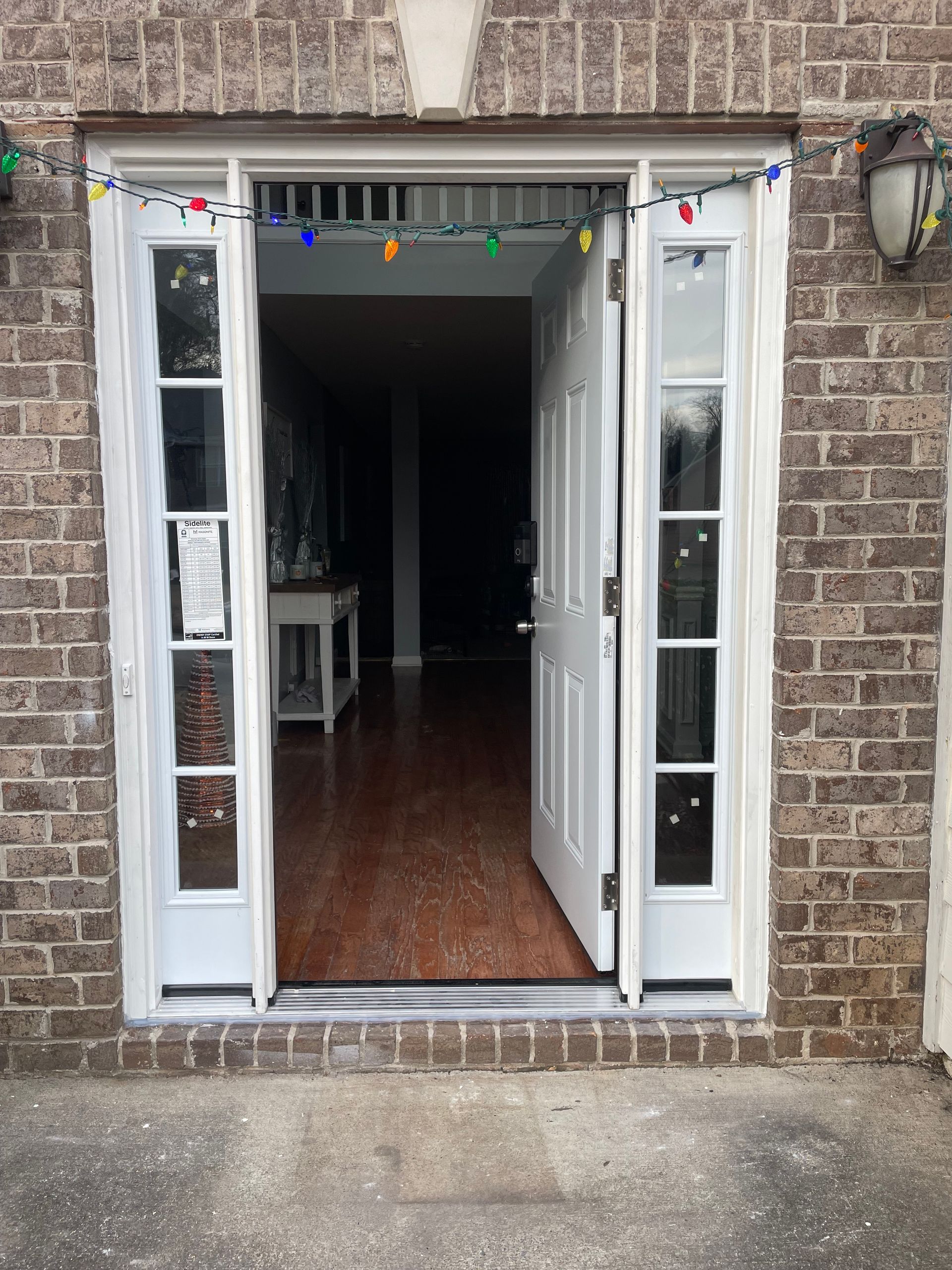 Open white front door with sidelights, colorful lights above, leading to interior with hardwood floors.