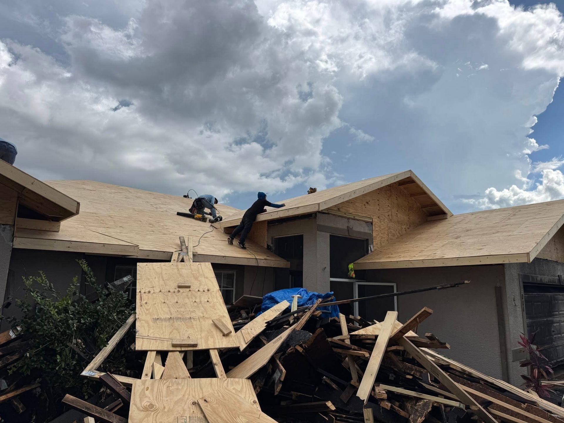 Two people installing plywood on a house roof under a cloudy sky.