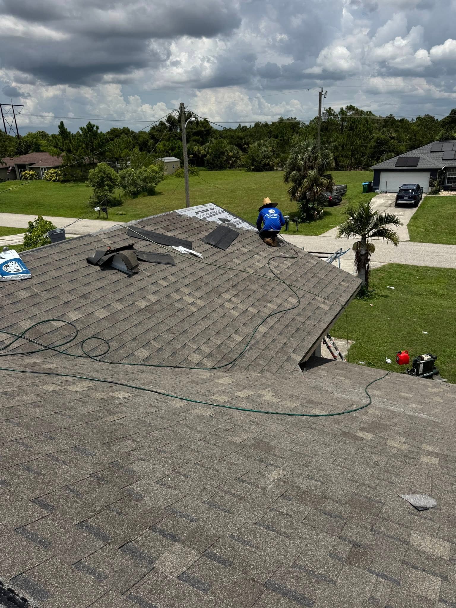 Person installing shingles on a house roof. Blue work clothes, materials, and suburban setting. 