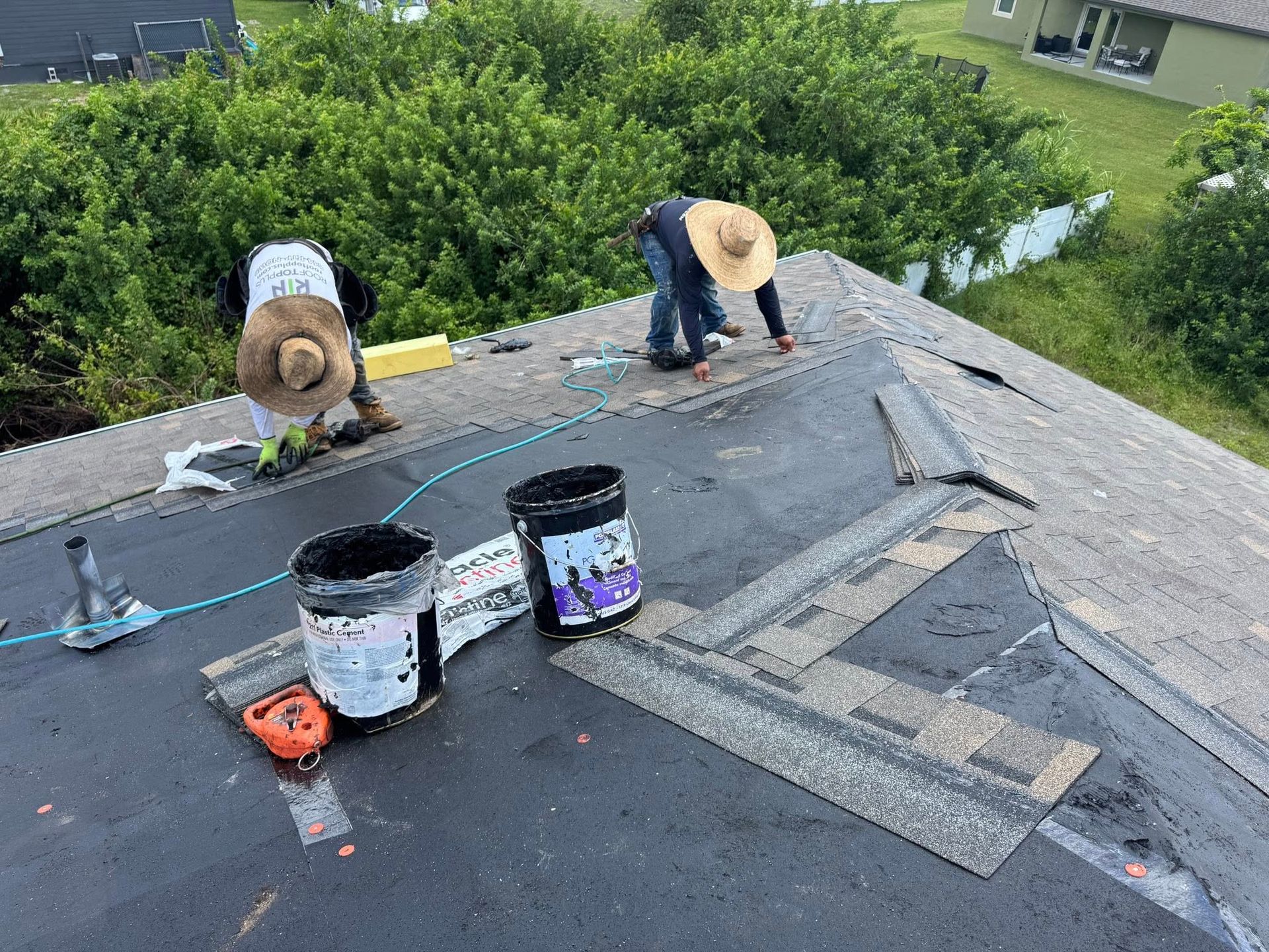 Two roofers in straw hats work on a dark shingle roof with buckets and tools.