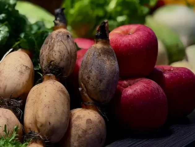 Close-up of fresh produce: lotus roots, red apples, green leafy vegetables, and a basket of oranges.