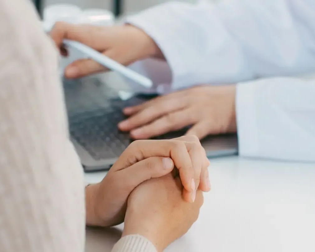 Doctor consulting with a patient at a desk, hands clasped, laptop open.