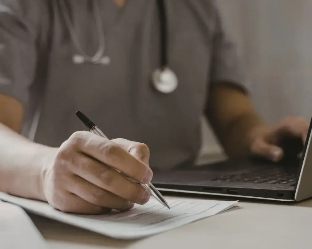 Person in scrubs, stethoscope, writes on paper, types on laptop.