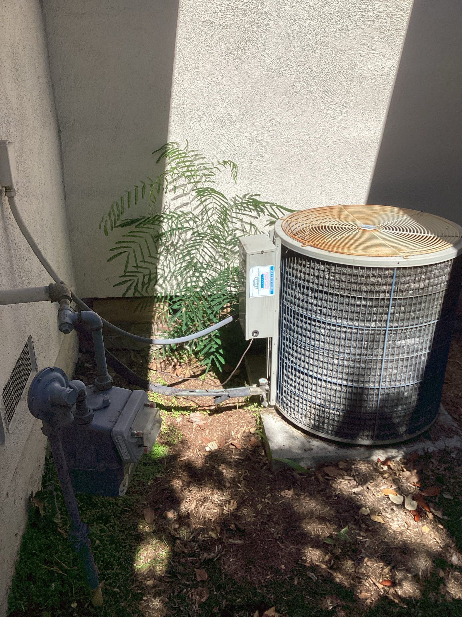 AC unit next to a building, with utility pipes and some plants.