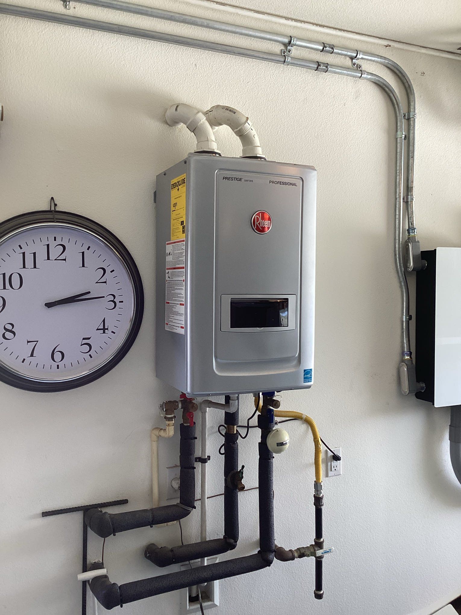 Tankless water heater installed on a white wall, with pipes, a clock, and electrical conduit.