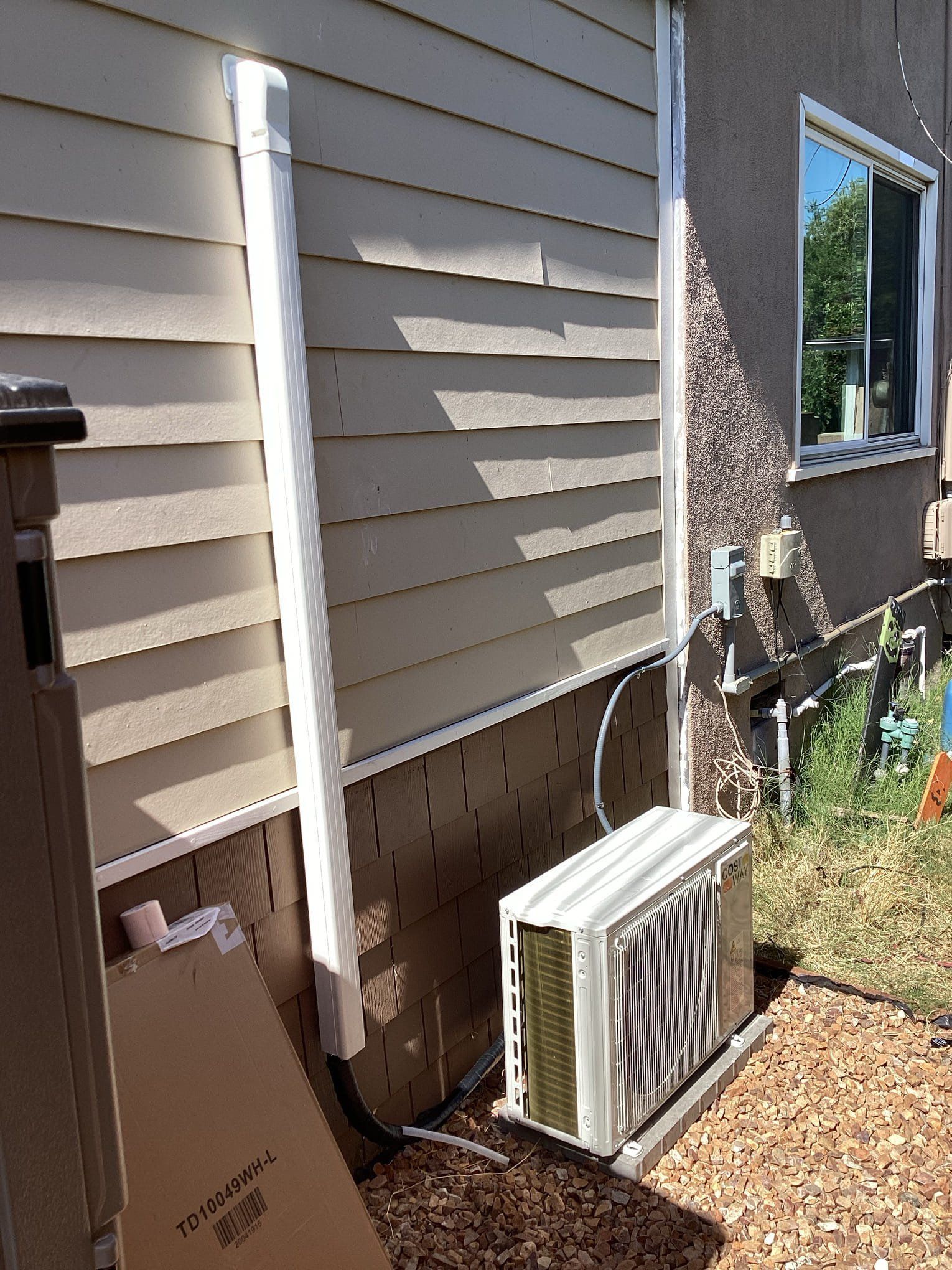 Exterior view of an air conditioning unit next to a house with beige siding, a window, and a white downspout.