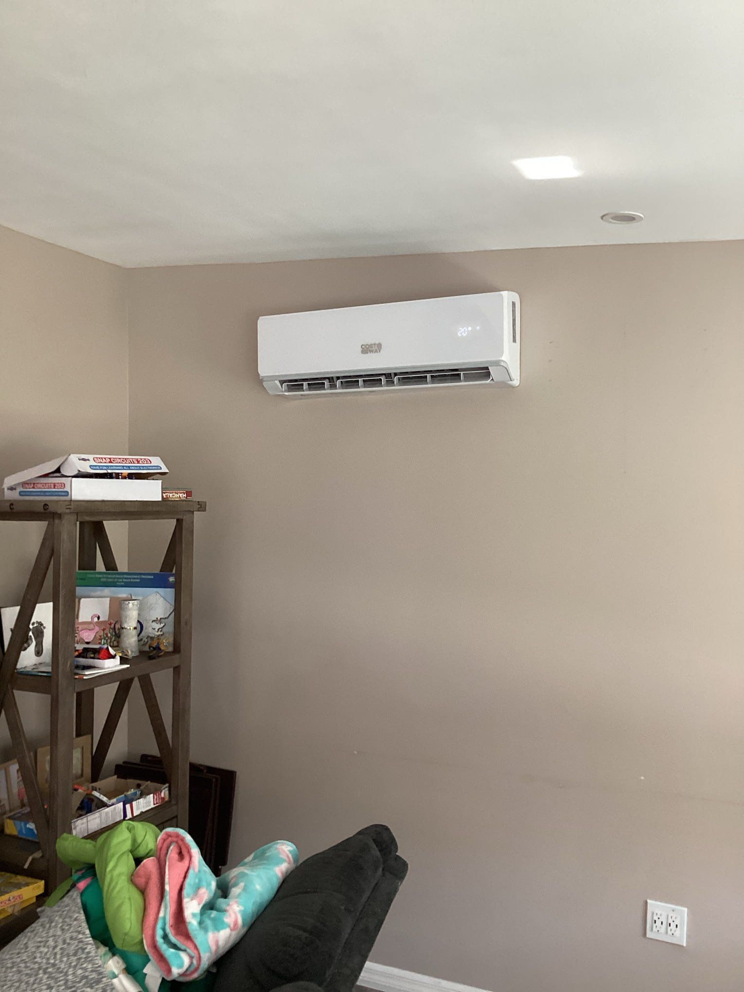 White wall-mounted air conditioner on beige wall. Wooden shelf with items, sofa, and electrical outlet.