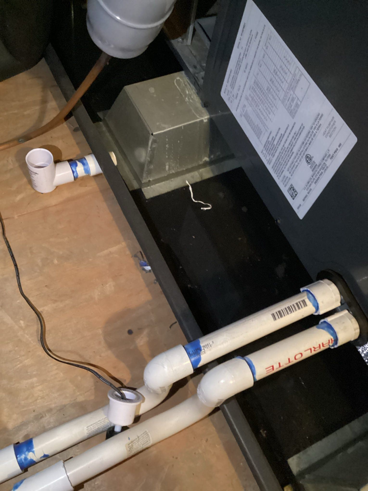 PVC pipes connected to a dark HVAC unit on a wooden floor, with a metal box and a label visible.
