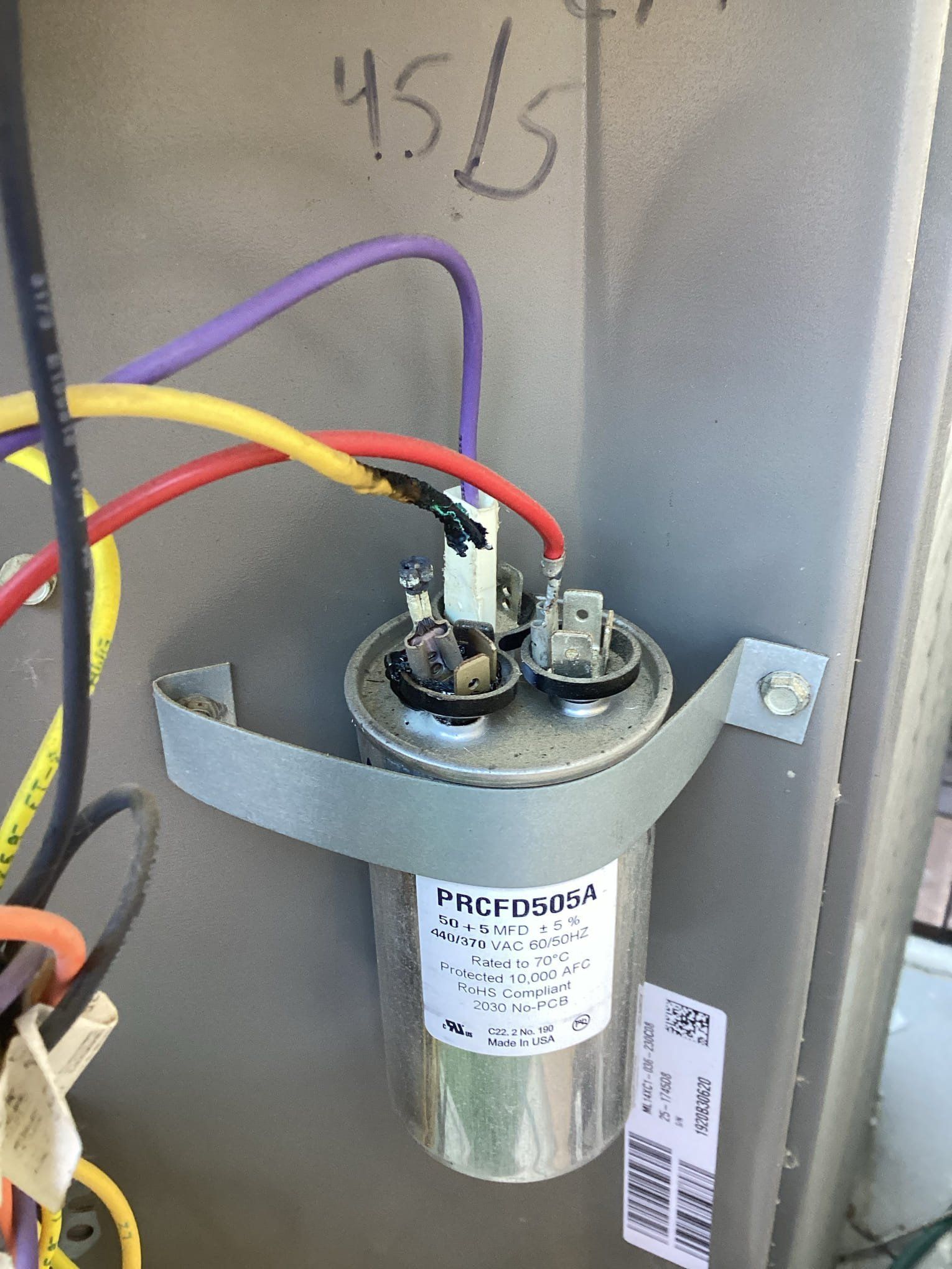 A capacitor, wires connected, mounted on an air conditioning unit. Wires are yellow, purple, and red.