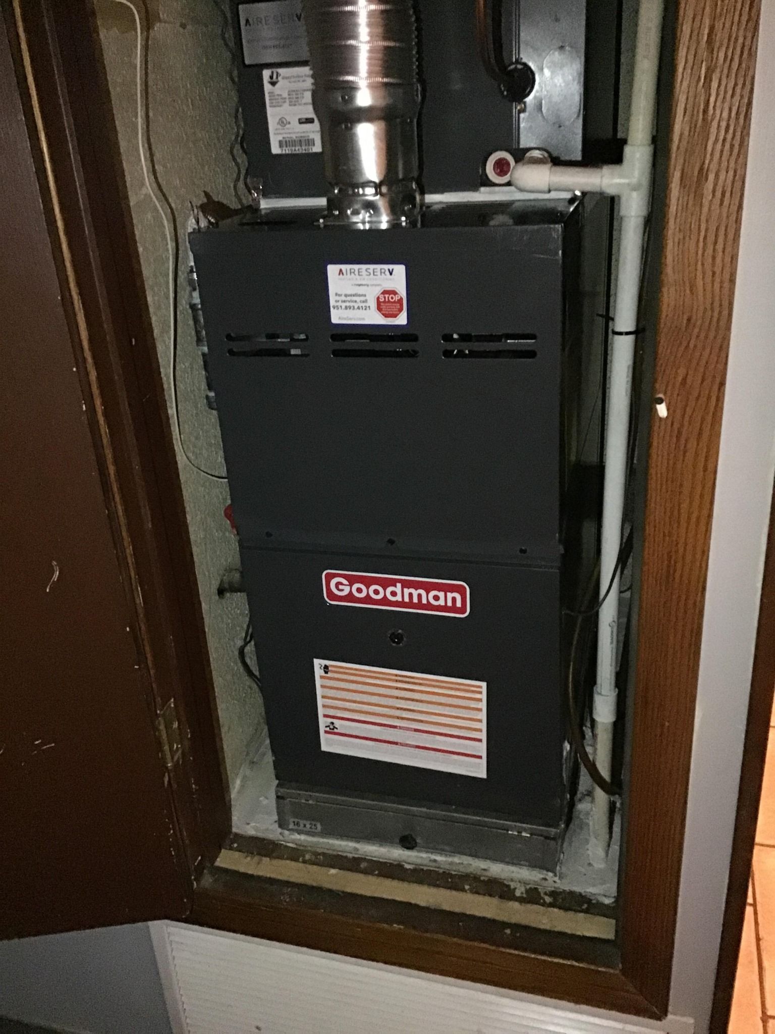 Goodman furnace in a closet. Black with silver flue, white pipes. Wooden door.