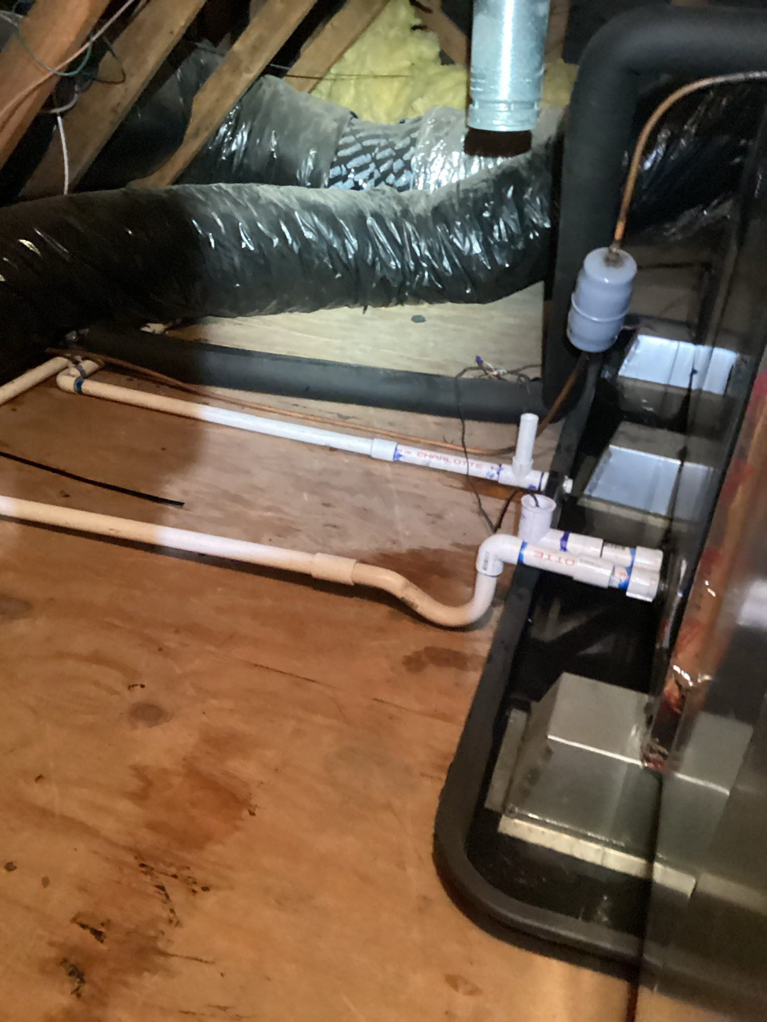 HVAC system components in attic: white PVC pipes, black insulation, and a float valve on a wooden floor.