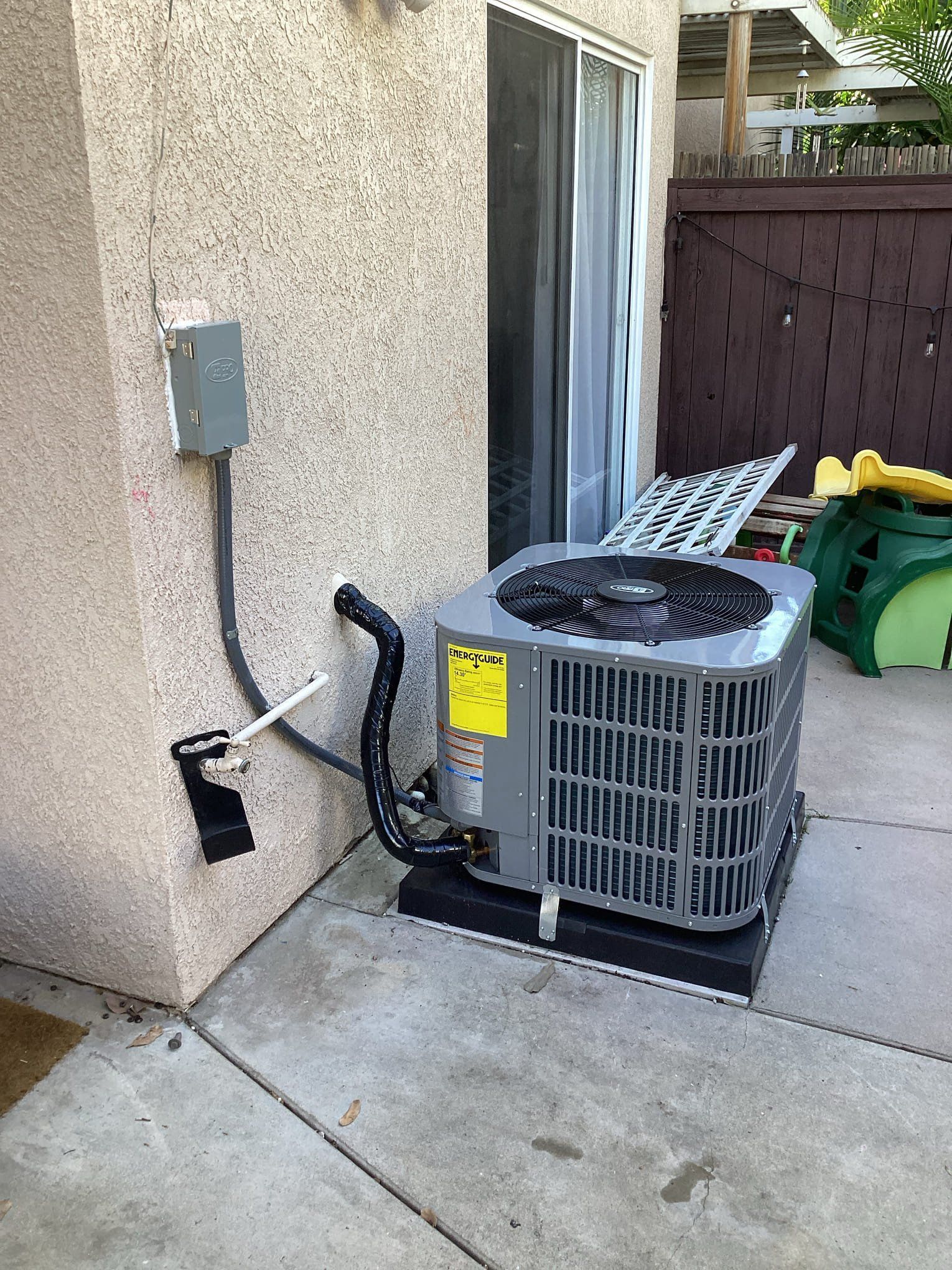 Outdoor air conditioning unit next to a stucco wall, with electrical box and sliding glass door.