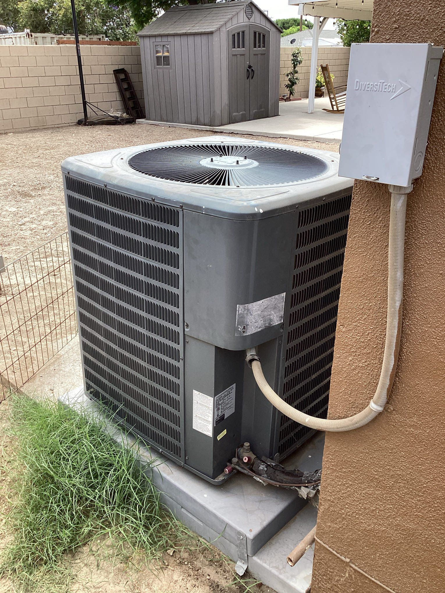 Air conditioning unit outdoors next to a building; a gray shed is in the background.