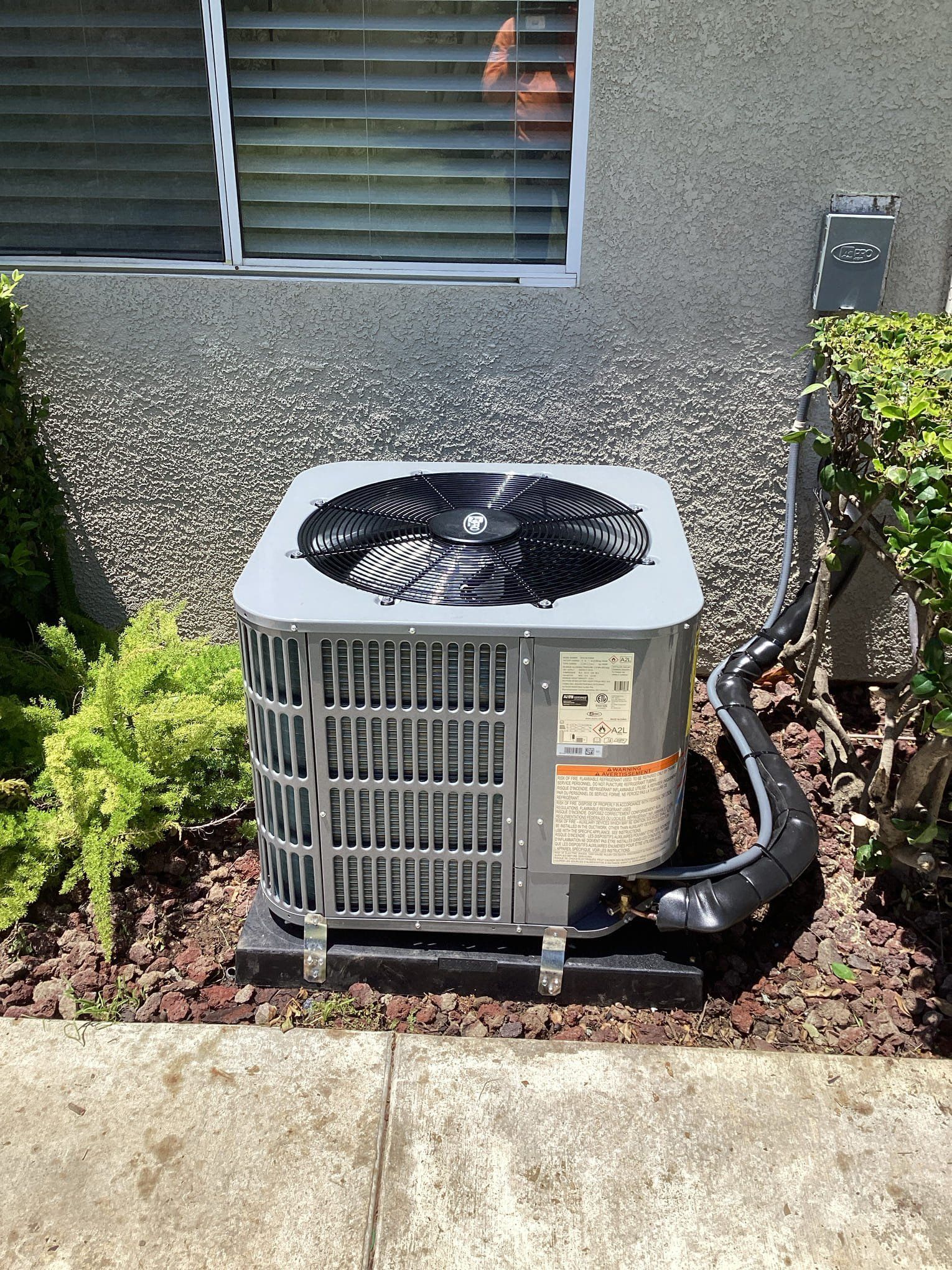 Exterior air conditioning unit on a dark base next to a bush and wall.