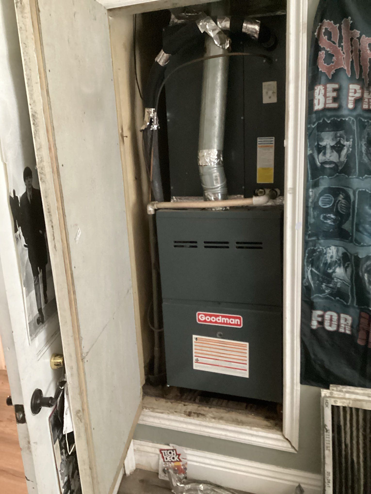 A furnace enclosed in a small closet; door open. The unit is grey with a red Goodman logo.