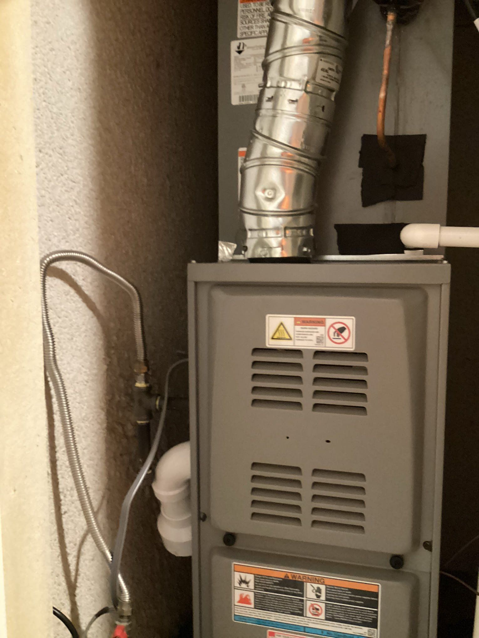 Furnace in a utility room with metal exhaust pipe, gas line, and warning labels.