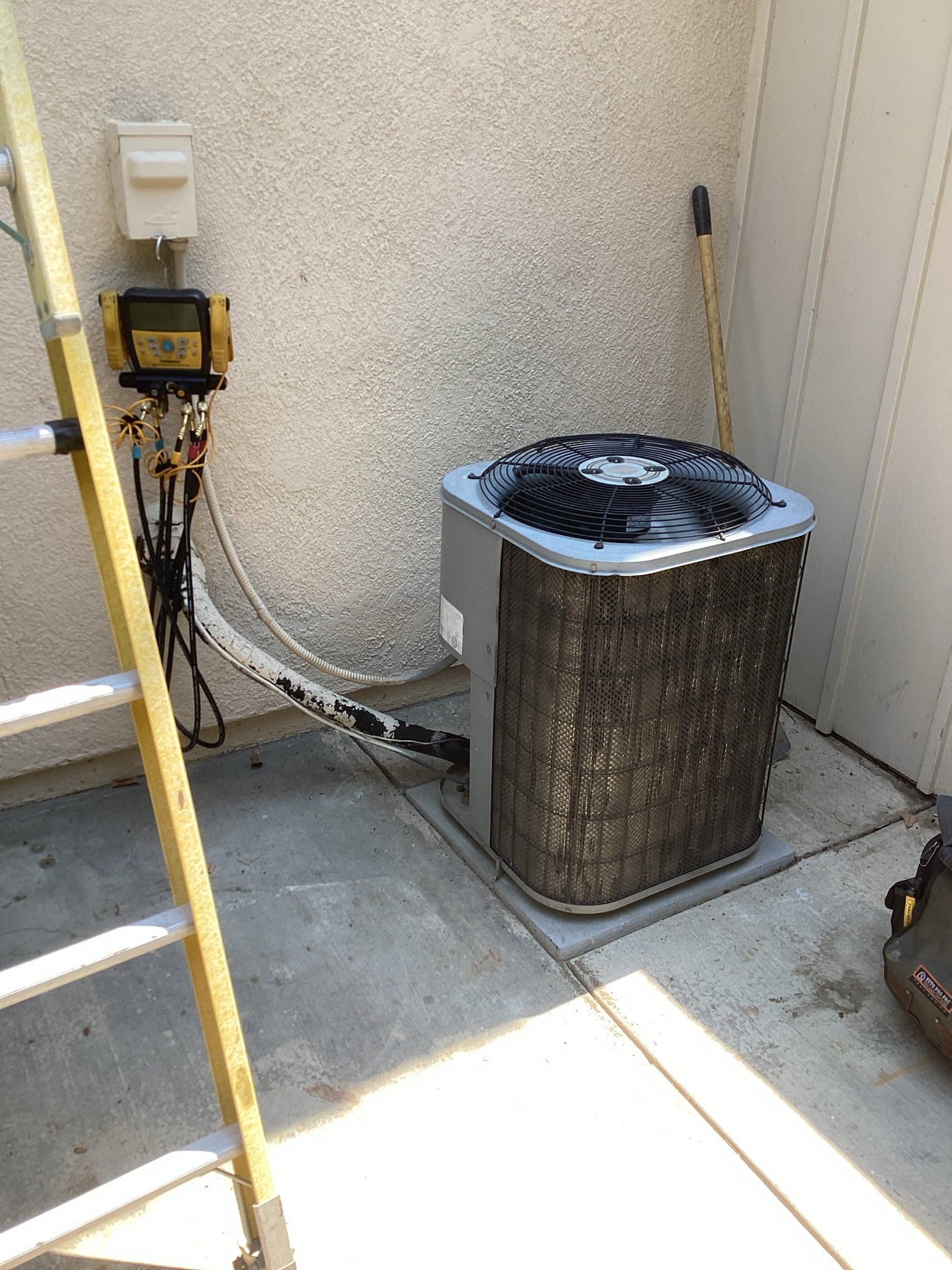 Outdoor air conditioning unit being serviced; ladder, gauges, and tool bag present.