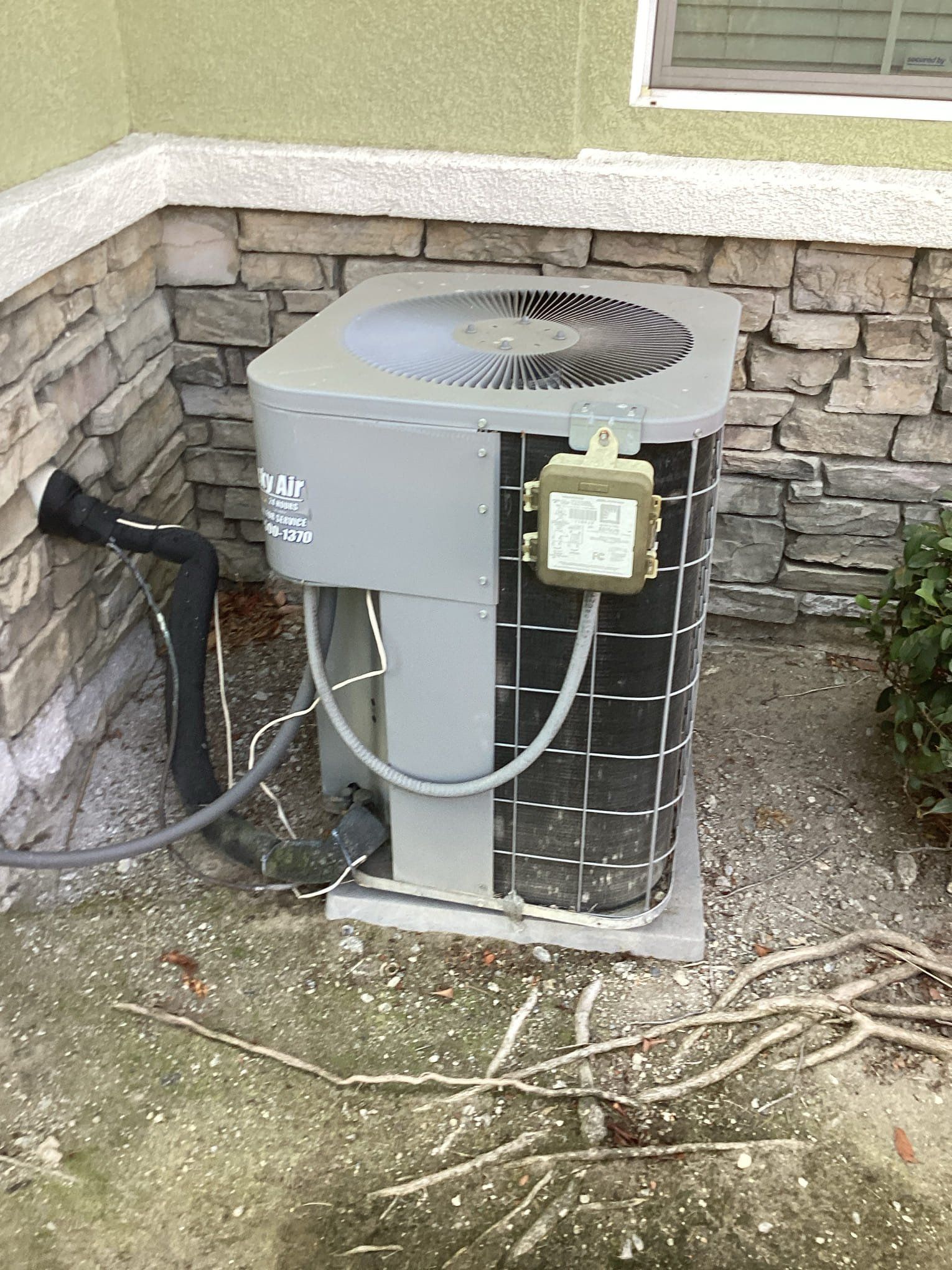 Air conditioning unit outside a building, gray with a protective cage, next to a stone wall.