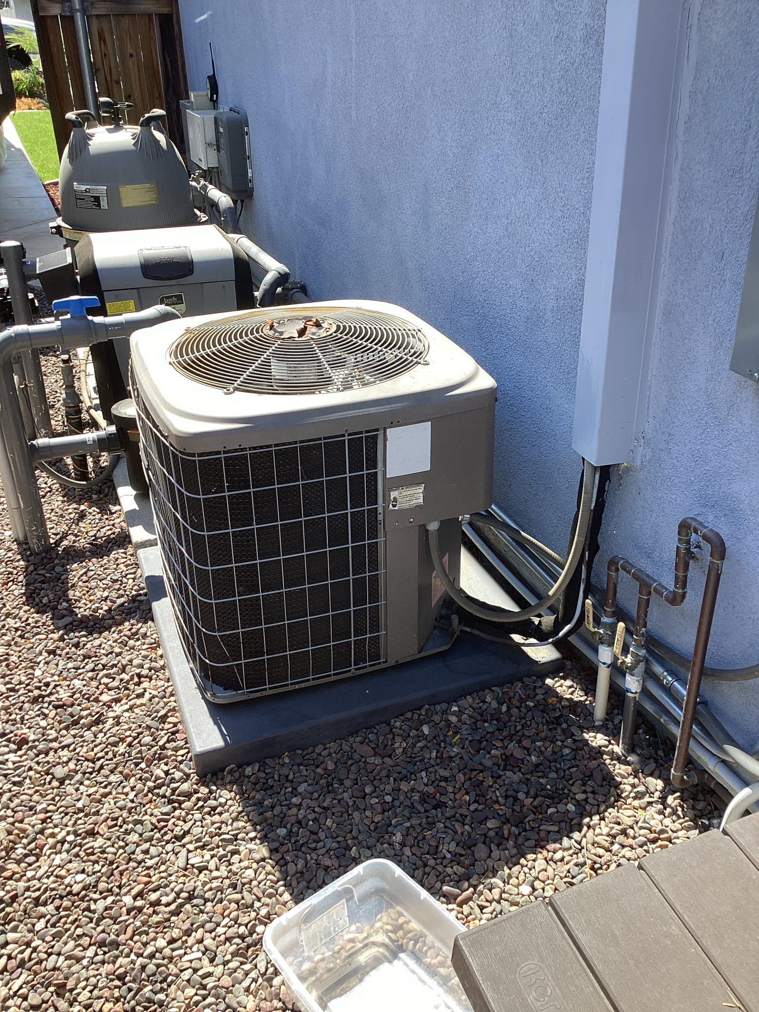 Air conditioning unit outside a house, on a black pad with gravel. Plumbing, pool equipment, and pipes are also present.