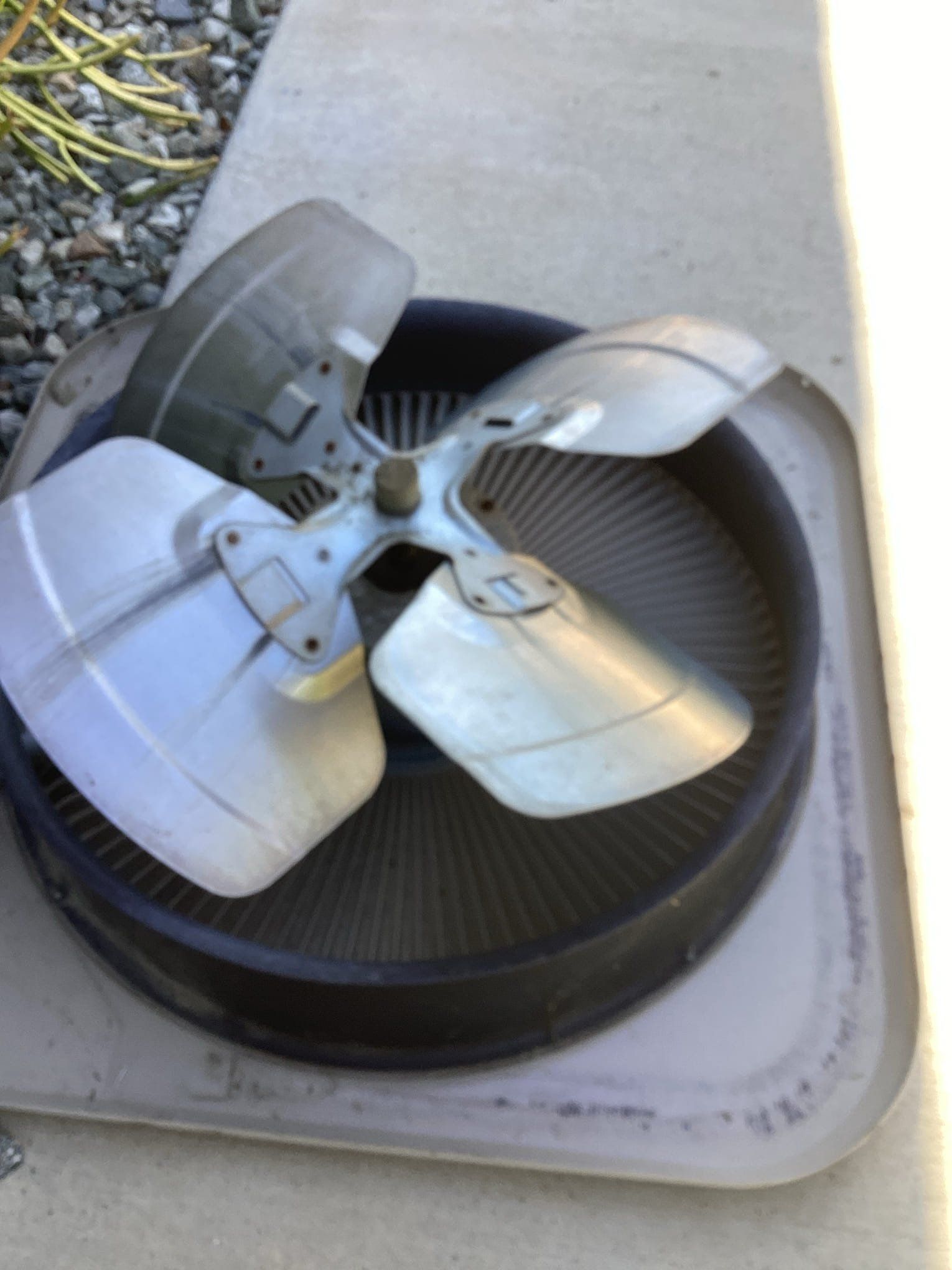 A damaged air conditioner fan with four silver blades, sitting outdoors on a white platform.