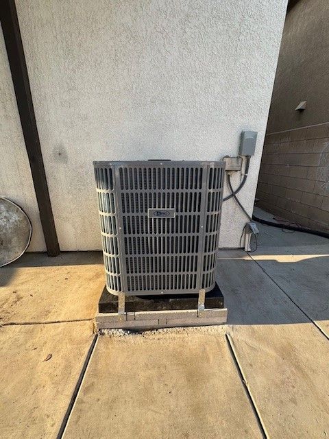 Air conditioning unit against a light wall, resting on a concrete pad. Electrical box on the wall.