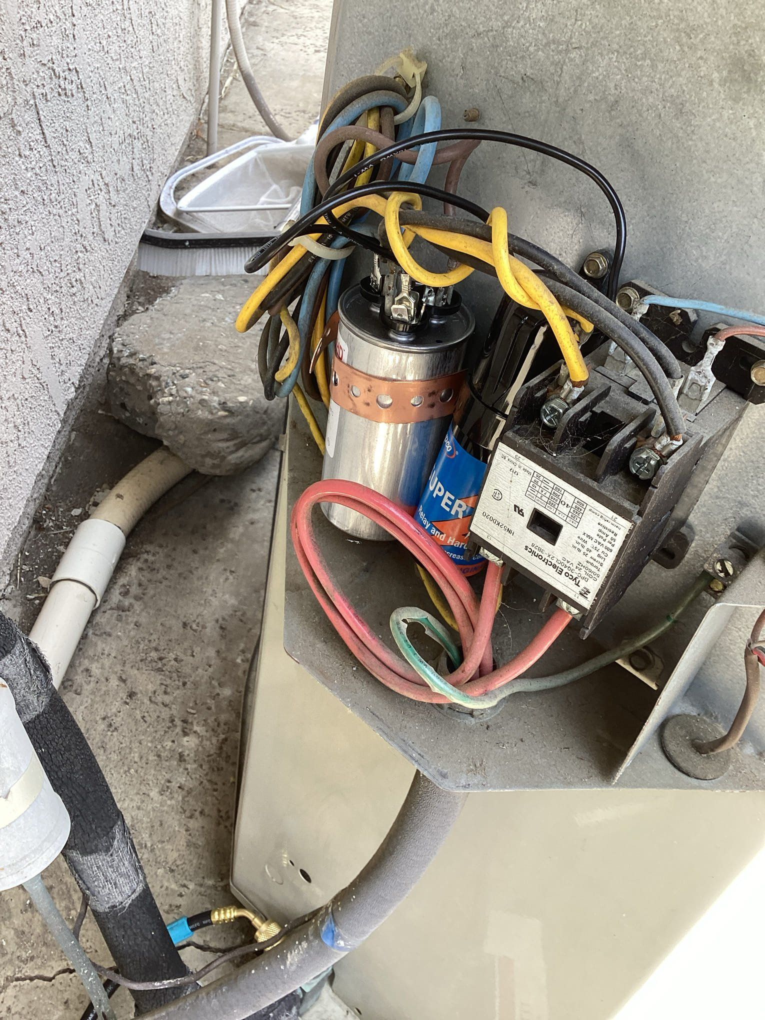 Air conditioning unit's electrical components: capacitor, contactor, and wiring against a concrete wall.
