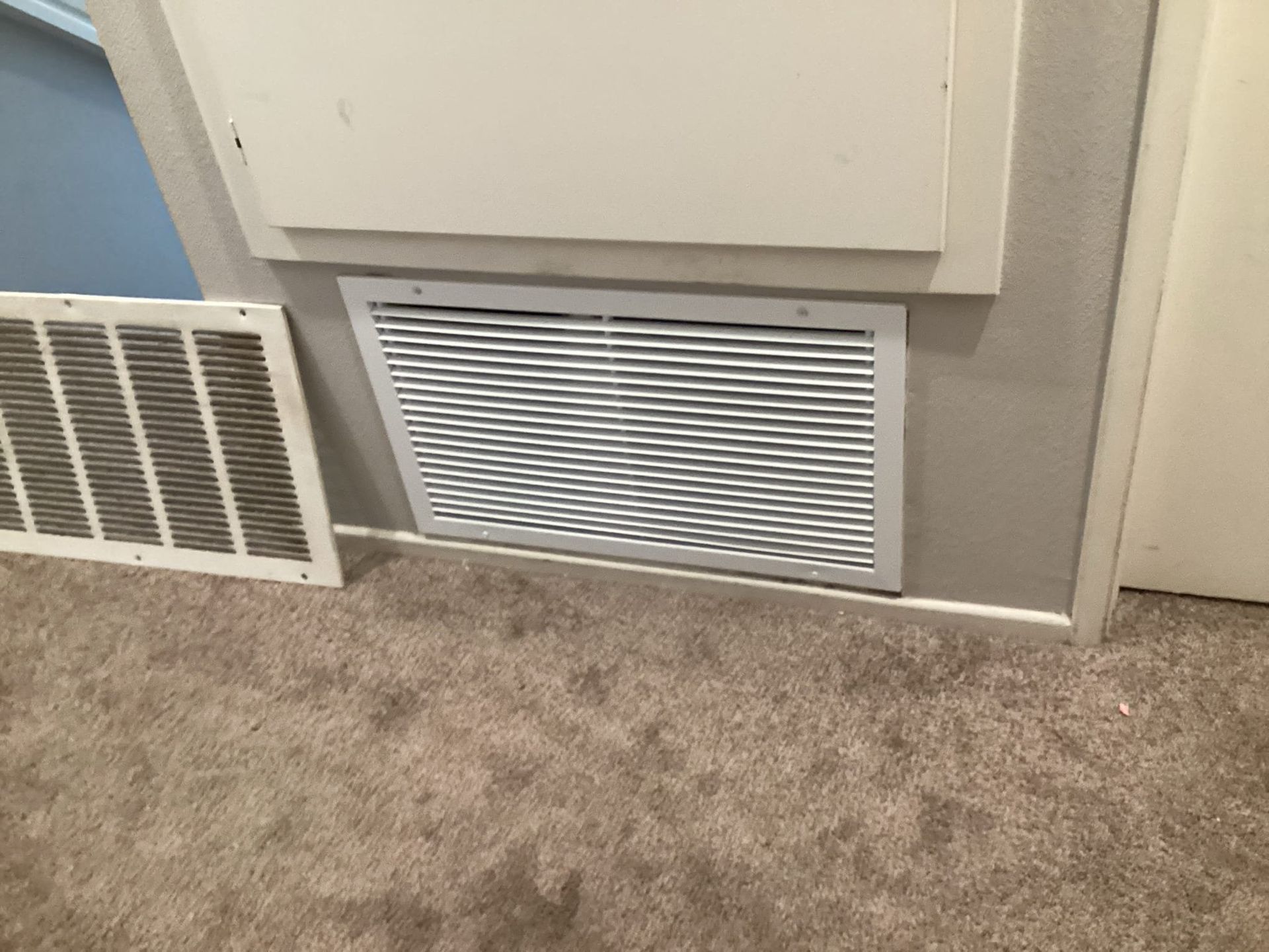 Ventilation grate on a wall above carpet. A removed air filter sits on the floor.