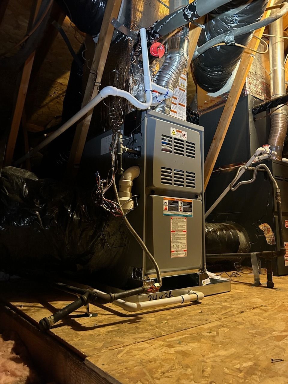 Furnace in an attic. Gray metal unit with attached ductwork, pipes, and wiring on wooden flooring.