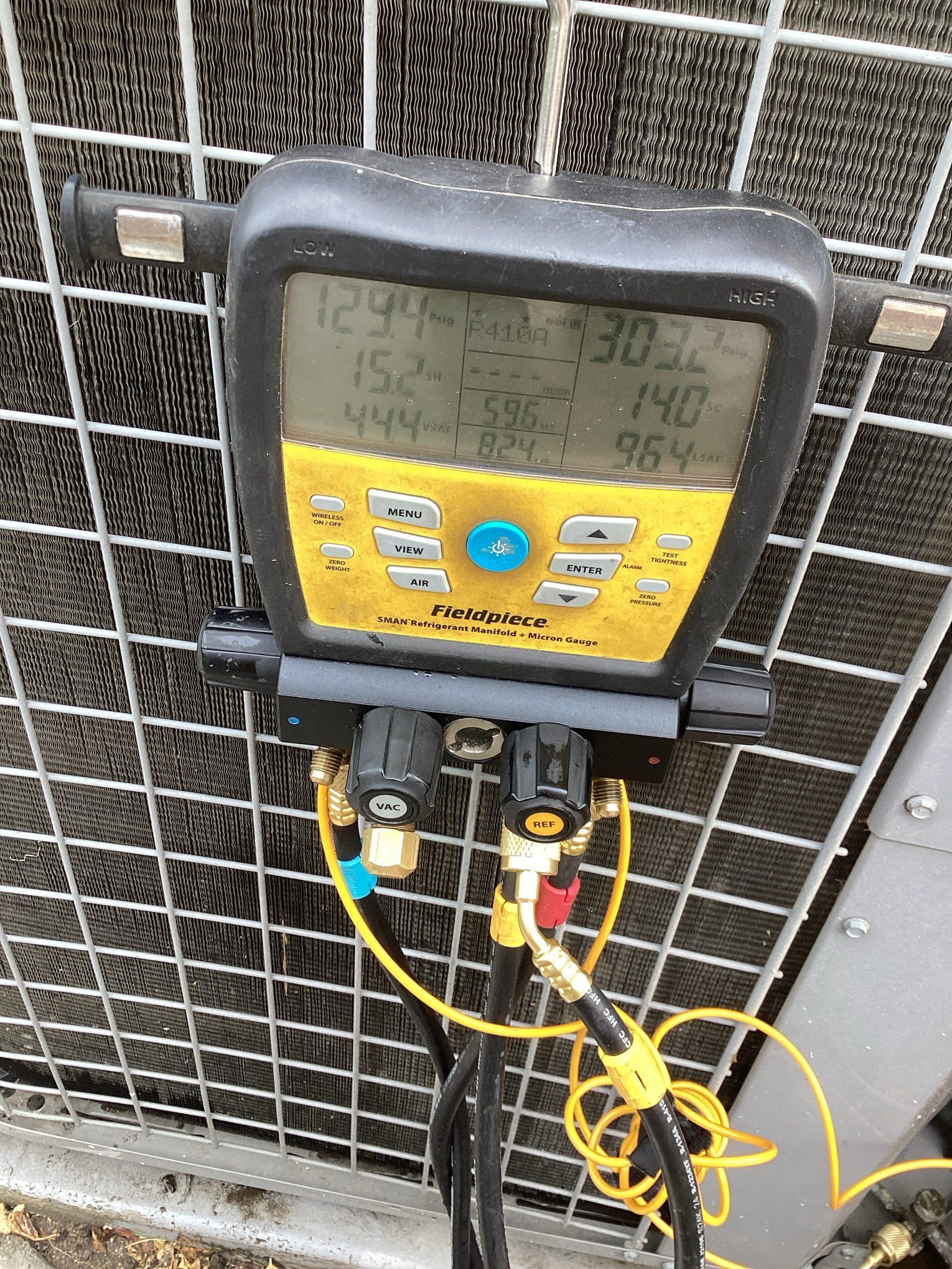 HVAC gauge display showing readings, connected to outdoor unit. Yellow hoses, black control valves, against a metal grate.