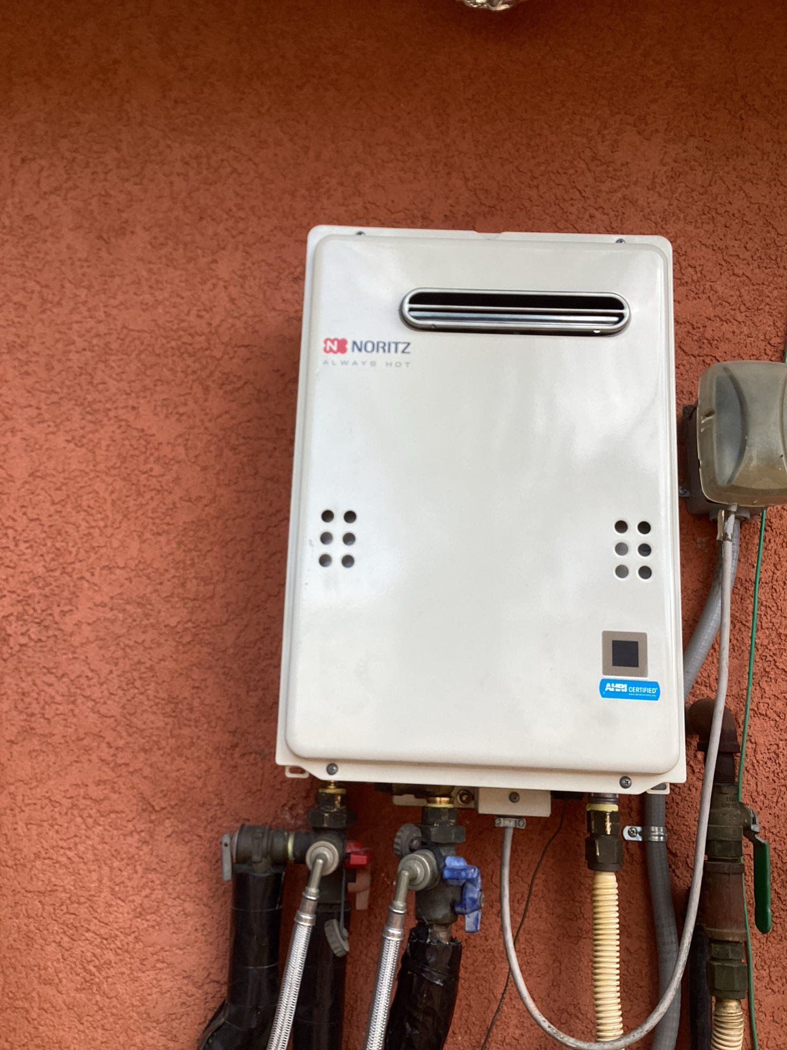 White tankless water heater on a red stucco wall, with pipes and connections.
