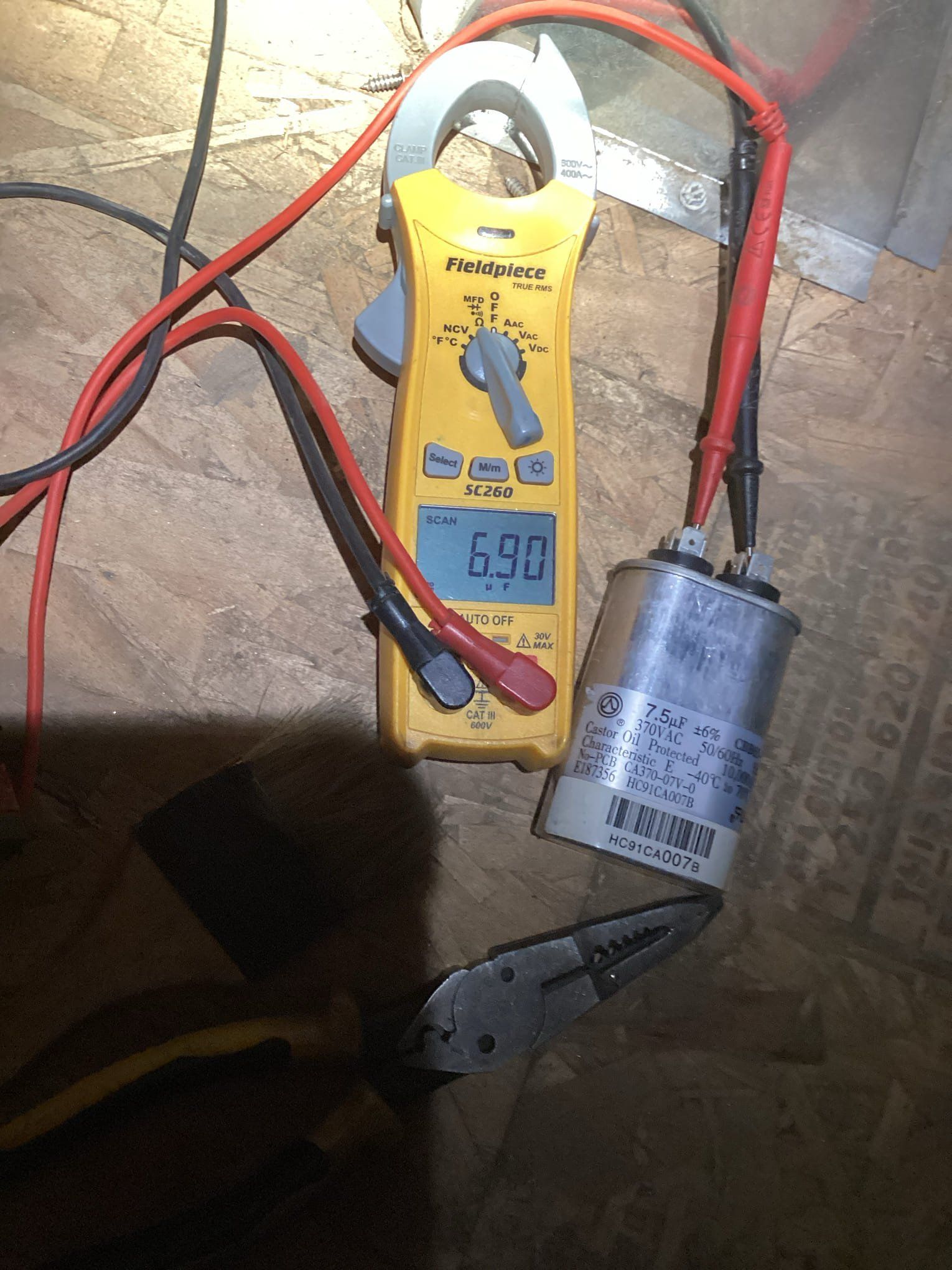 Yellow multimeter displaying 6.90 amps, clamped around a wire connected to an air conditioning capacitor.