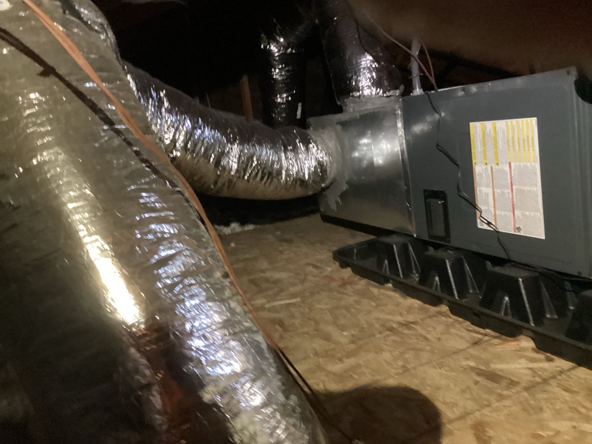 HVAC unit and ductwork in an attic setting. Silver insulated ducts connect to a gray unit on a black base.