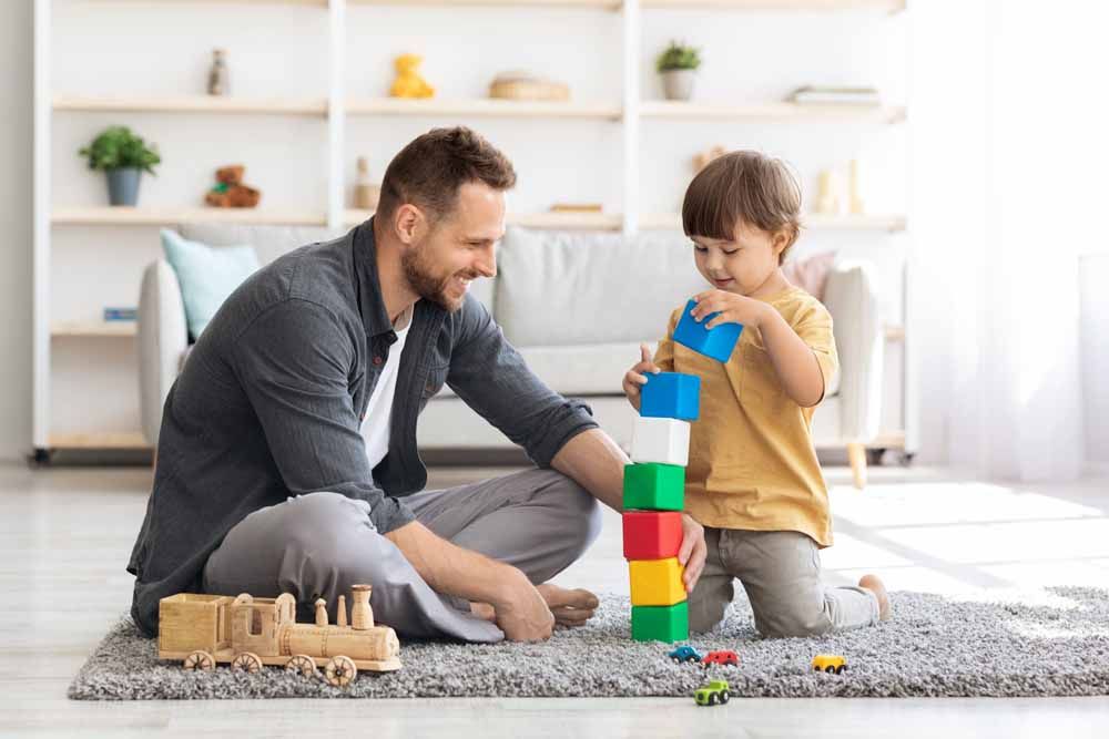 Father and son building a tower of colorful blocks together on a rug, smiling in a living room.